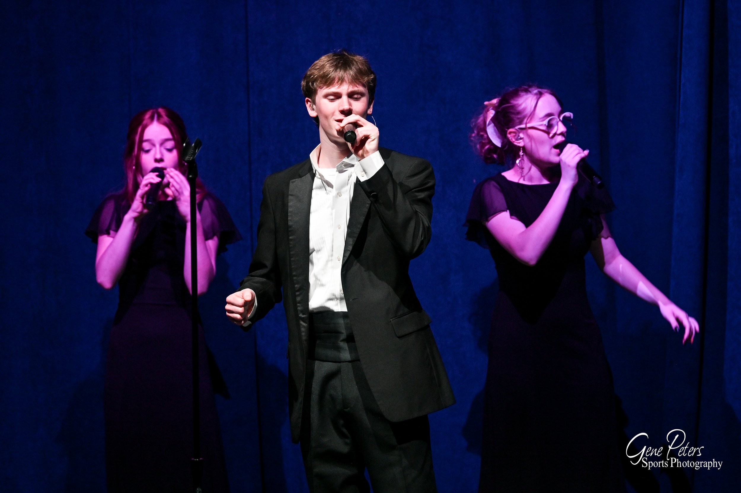 Three young performers singing on stage, with two girls in black dresses and a boy in a suit, against a dark blue curtain background.