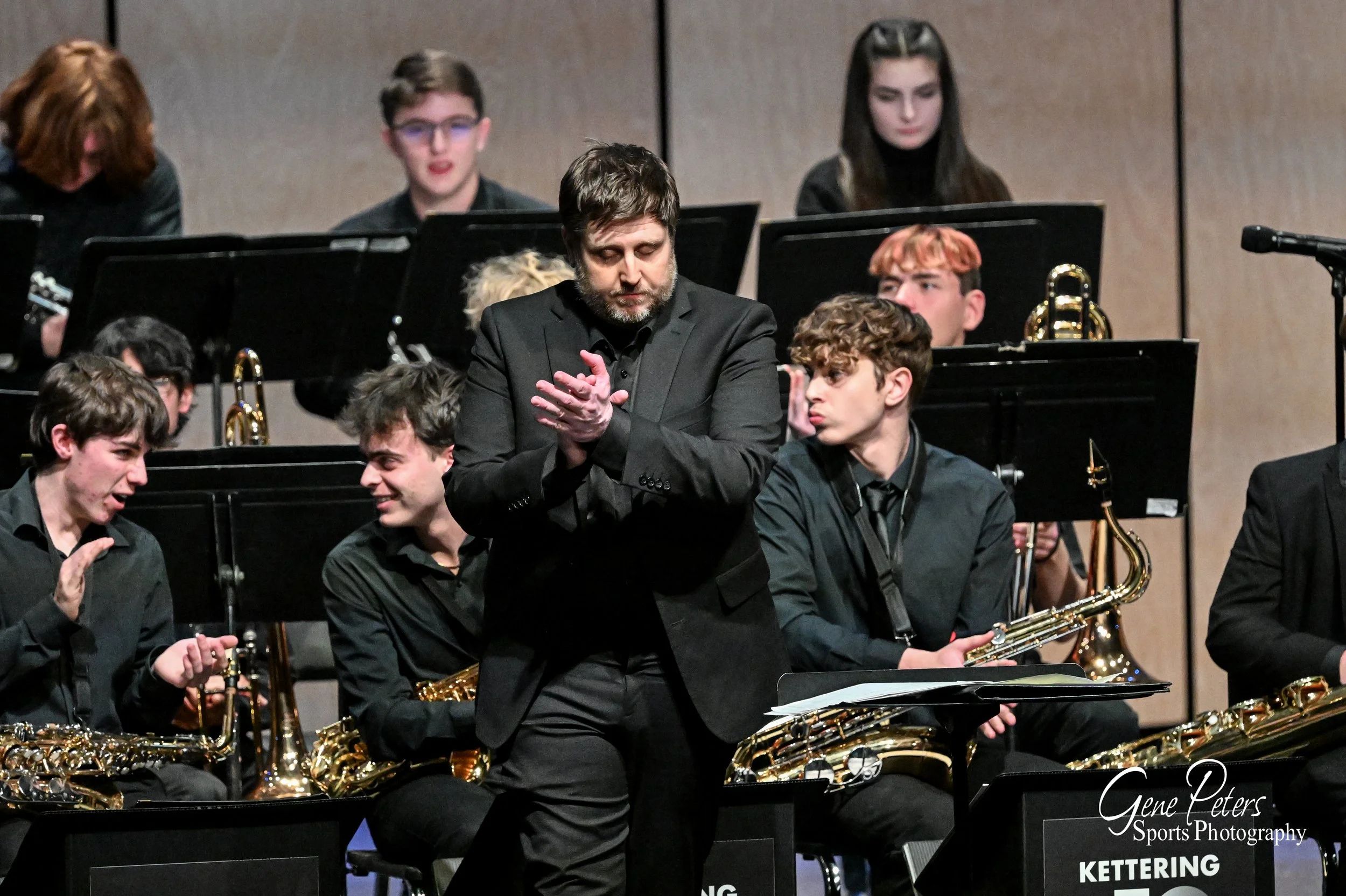 A group of young musicians in black attire, seated with brass and woodwind instruments, after a performance with a conductor in the center clapping. The scene is set in a concert hall with a wooden backdrop.