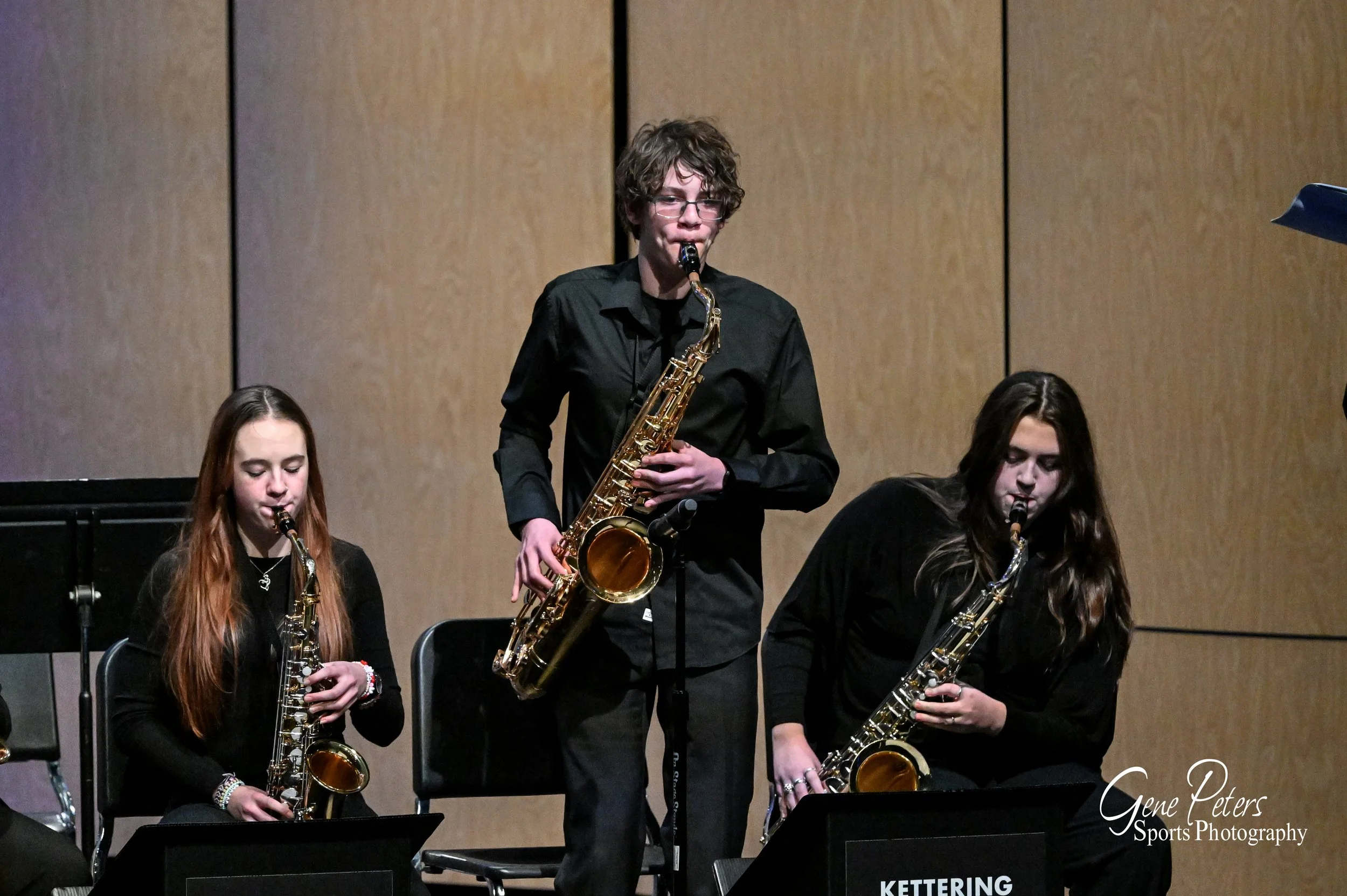 Three young musicians playing altosaxophones on stage during a performance, with a plain wooden backdrop behind them.
