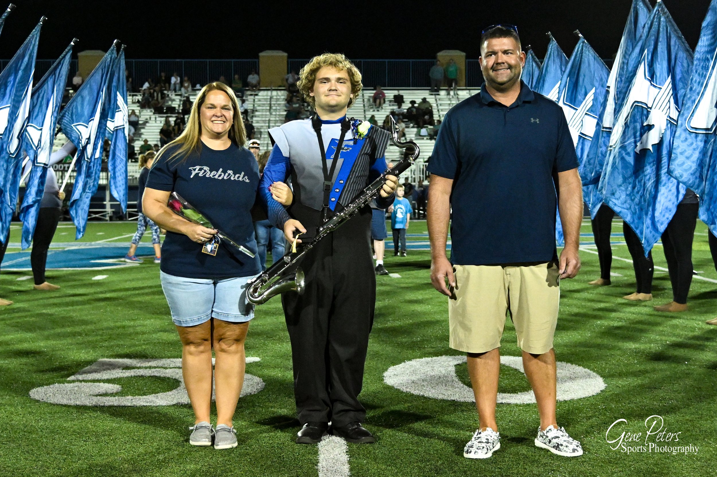 Three people standing on a football field at night with a band in the background. The person in the middle is a young man holding a clarinet, wearing a uniform, with a medal around his neck. The woman on the left is holding a bouquet of flowers. The 