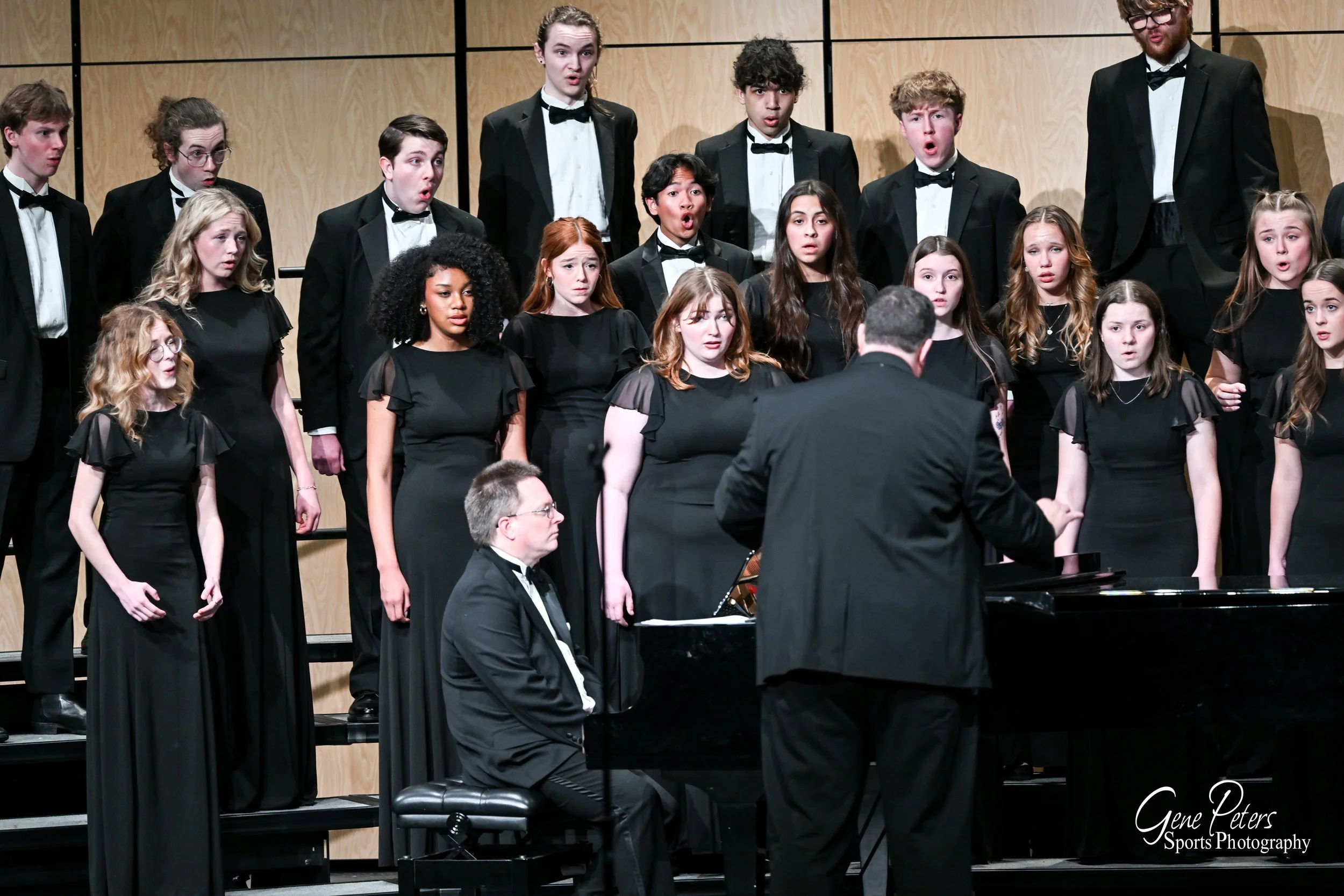 A choir of young men and women dressed in black, performing on stage with a conductor and pianist.