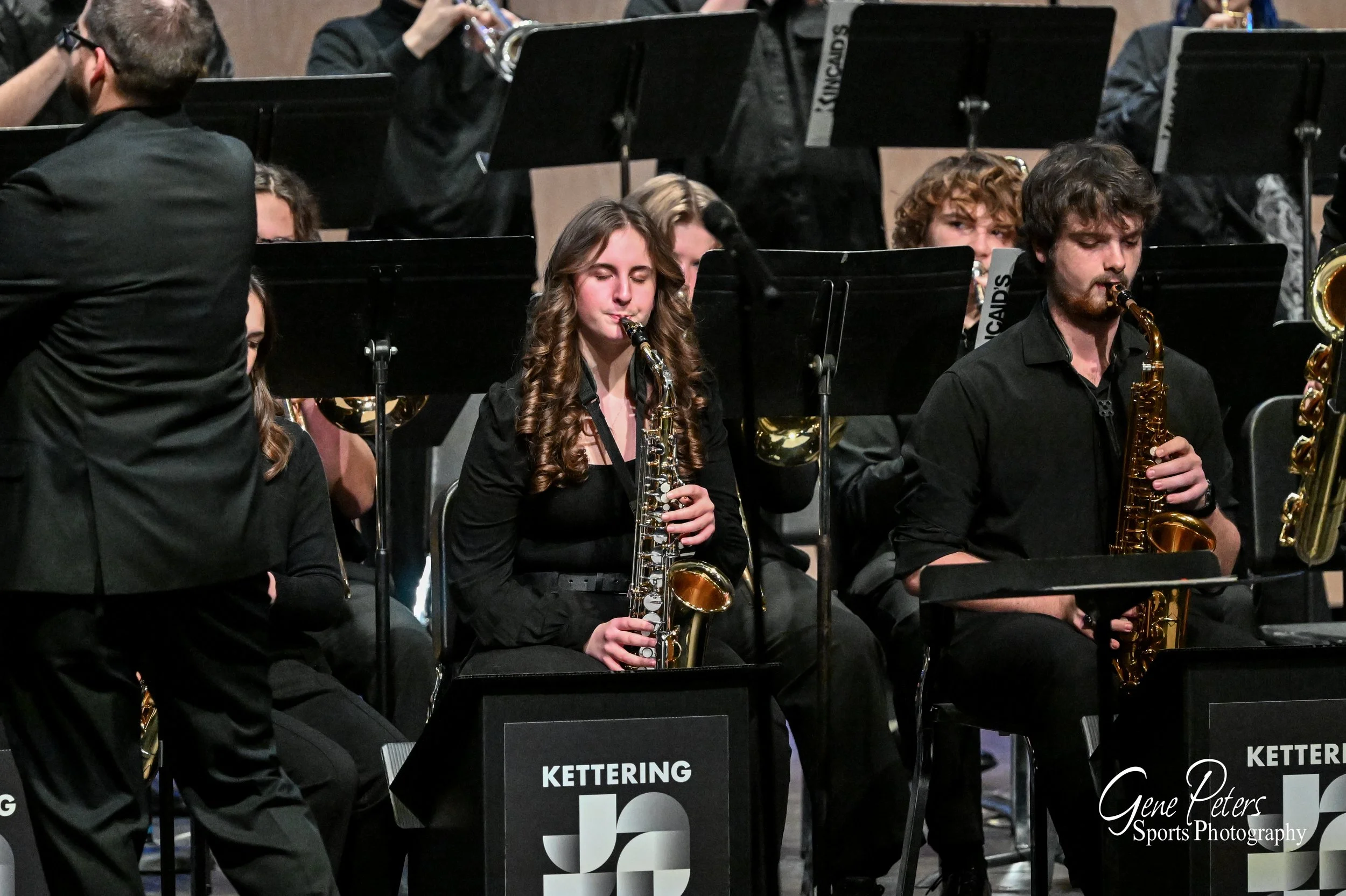 Musicians in an orchestra, including a young woman playing the clarinet and a young man playing the saxophone, seated behind black music stands with 'Kettering' written on them.