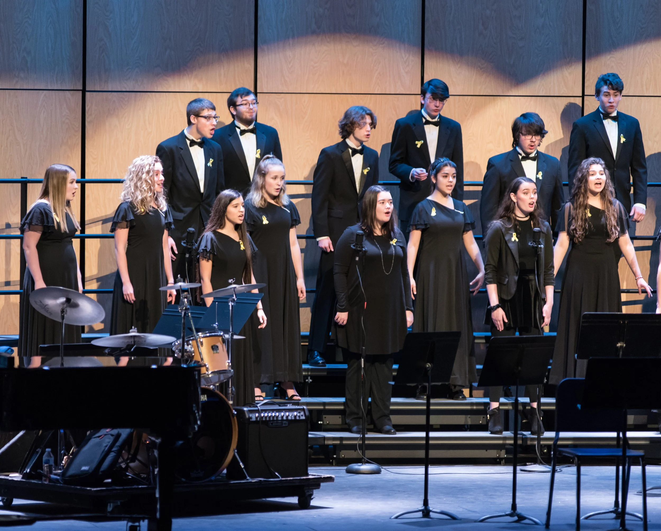 A choir of young men and women singing on stage during a performance, dressed in formal black attire with yellow ribbons, with musical instruments and microphones in front.