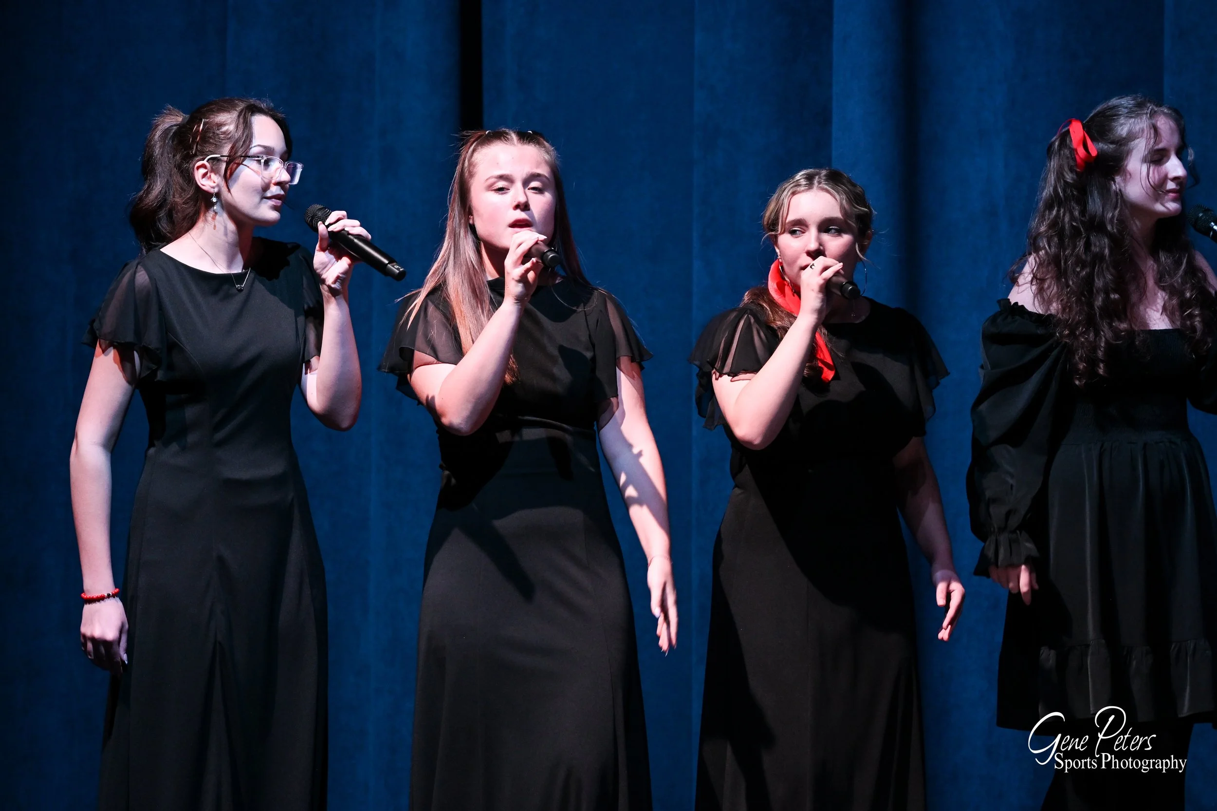 Four young women singing on stage with microphones, dressed in black outfits, with blue curtains in the background.