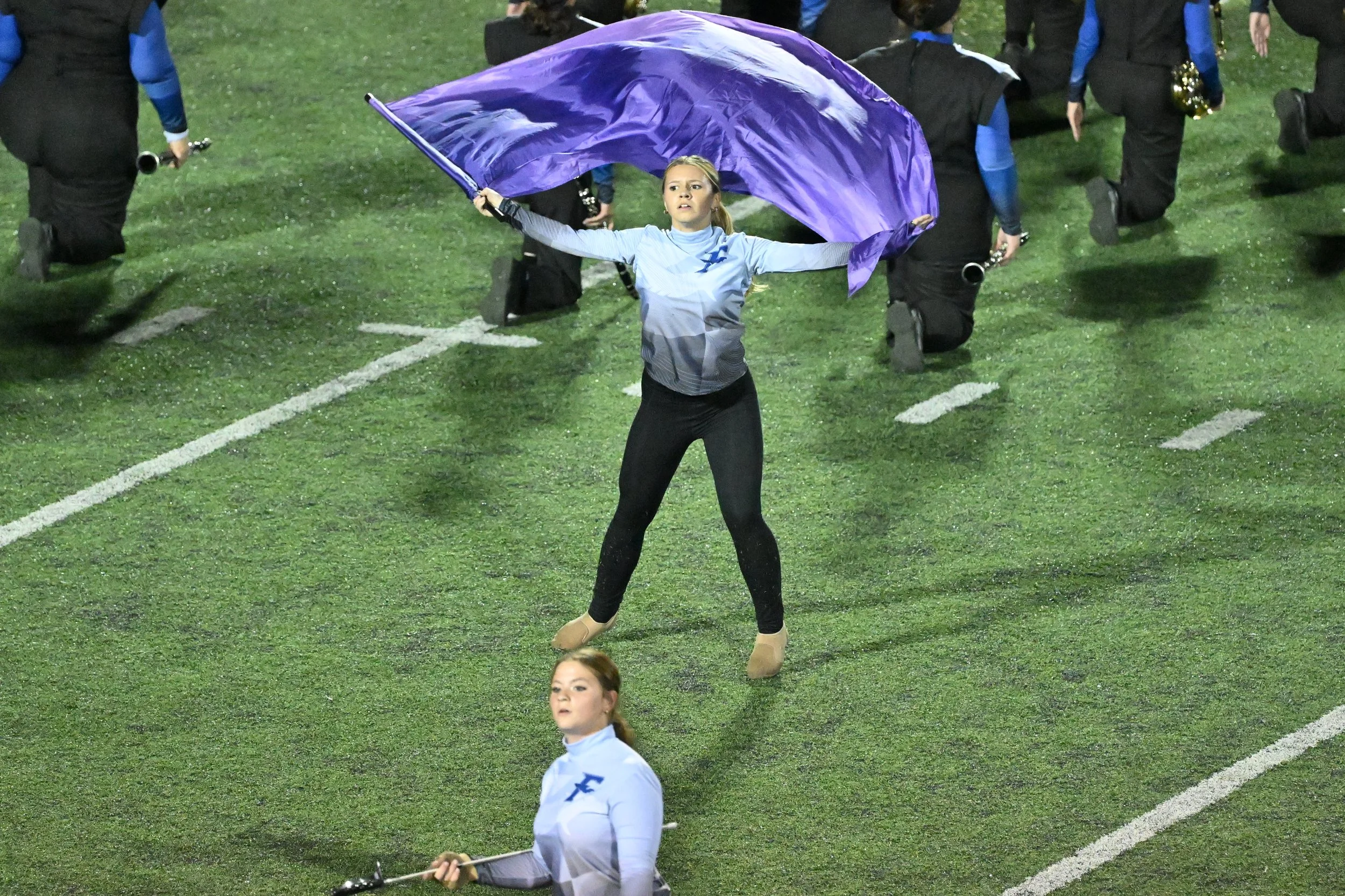 Young woman holding a purple flag, leading a marching band on a football field at night.