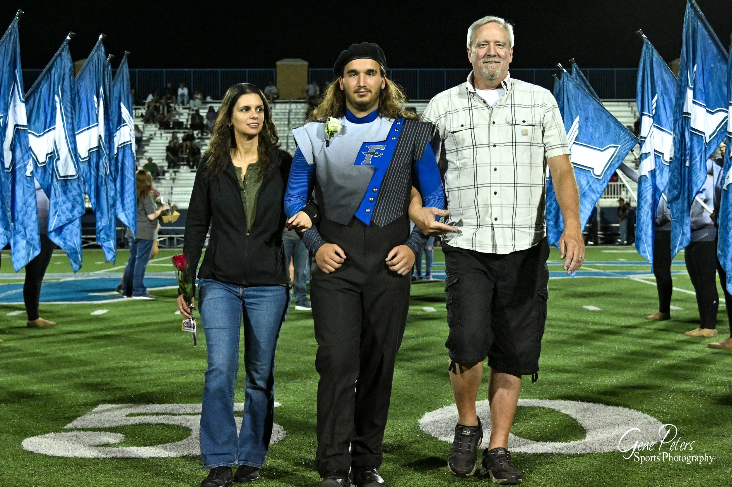 A young man in a football uniform walking on a field with his parents on either side, holding a flower, with blue flags and bleachers in the background.