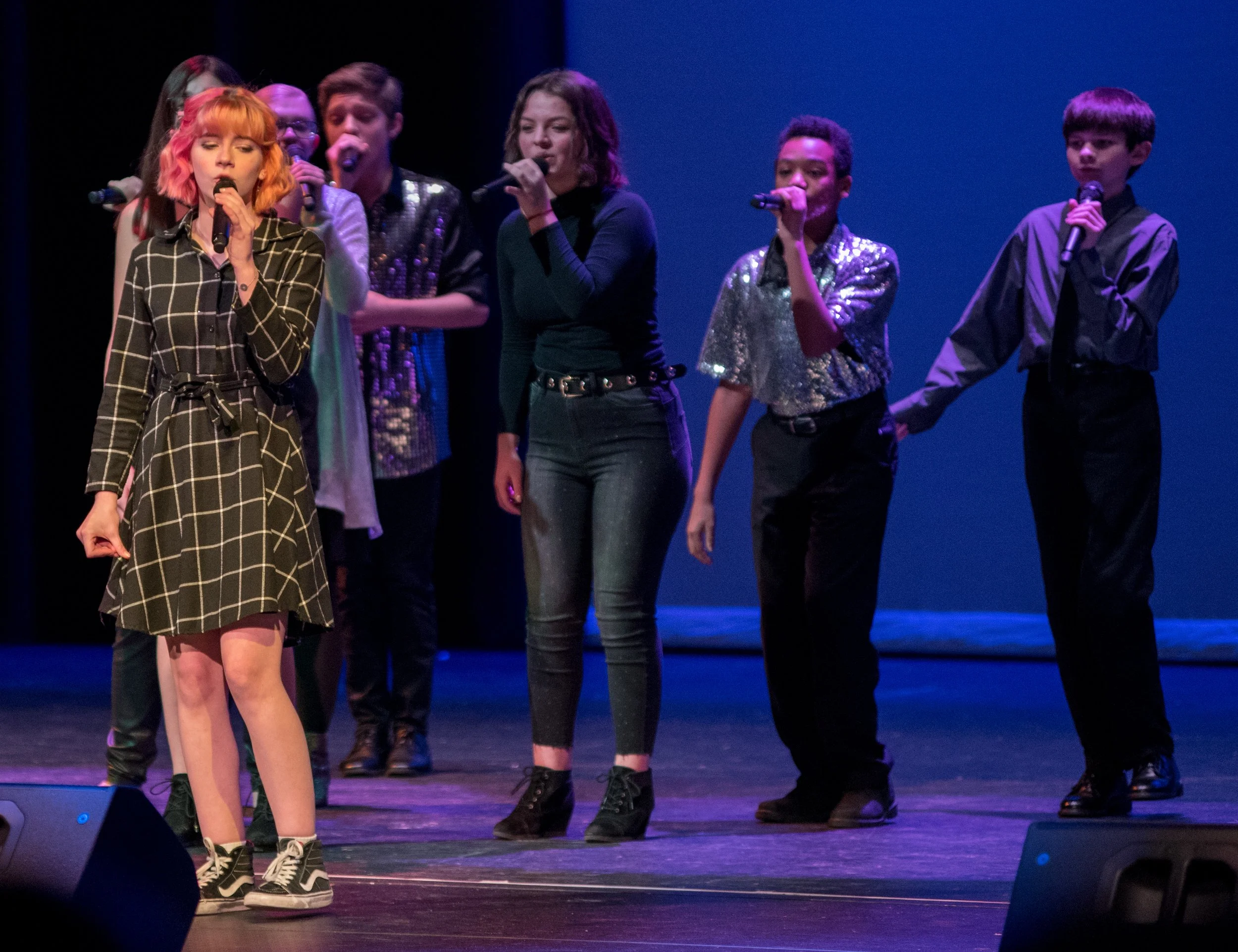 Group of young performers singing into microphones on stage, some with sequined outfits, with blue backdrop.