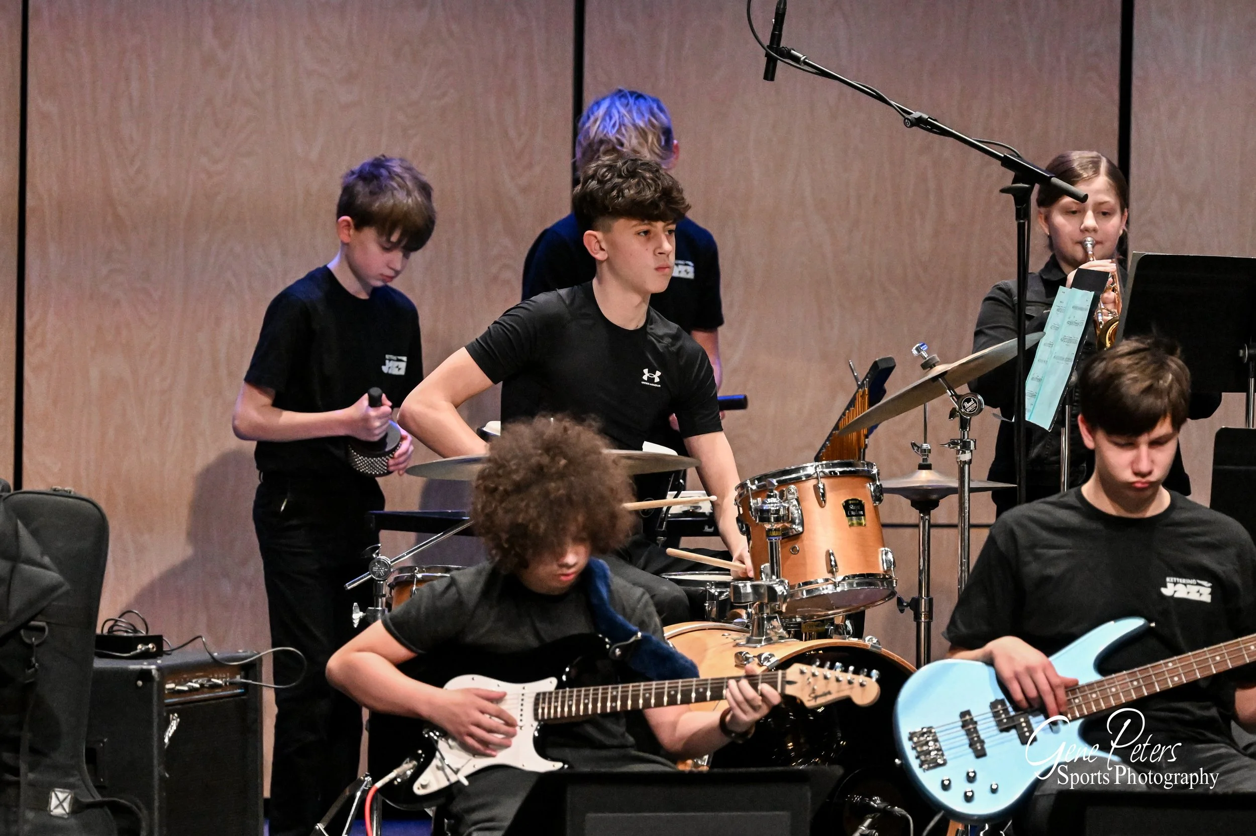 Group of young musicians performing on stage, playing various instruments including guitar, drums, and trumpet, under a wooden backdrop.