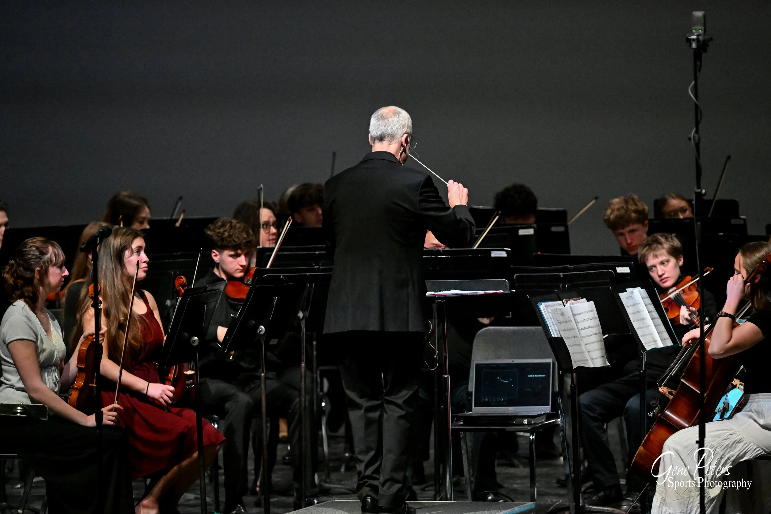 Conductor leading a youth orchestra during a performance, with musicians playing violins and cellos, and music stands with sheet music