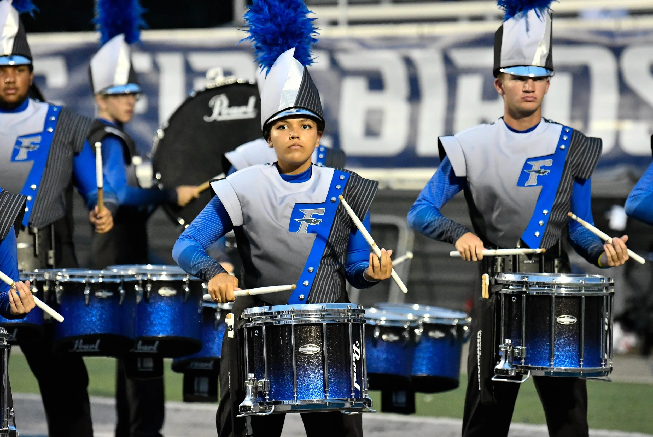 A marching band performing on a football field, with young musicians in blue and gray uniforms playing drums, some wearing tall hats with blue feathers.