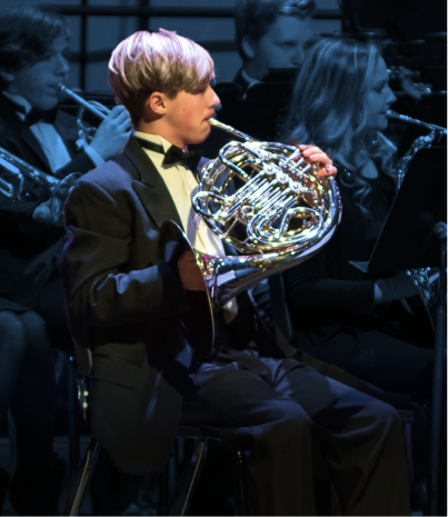 Young boy in formal attire playing a French horn in an orchestra.