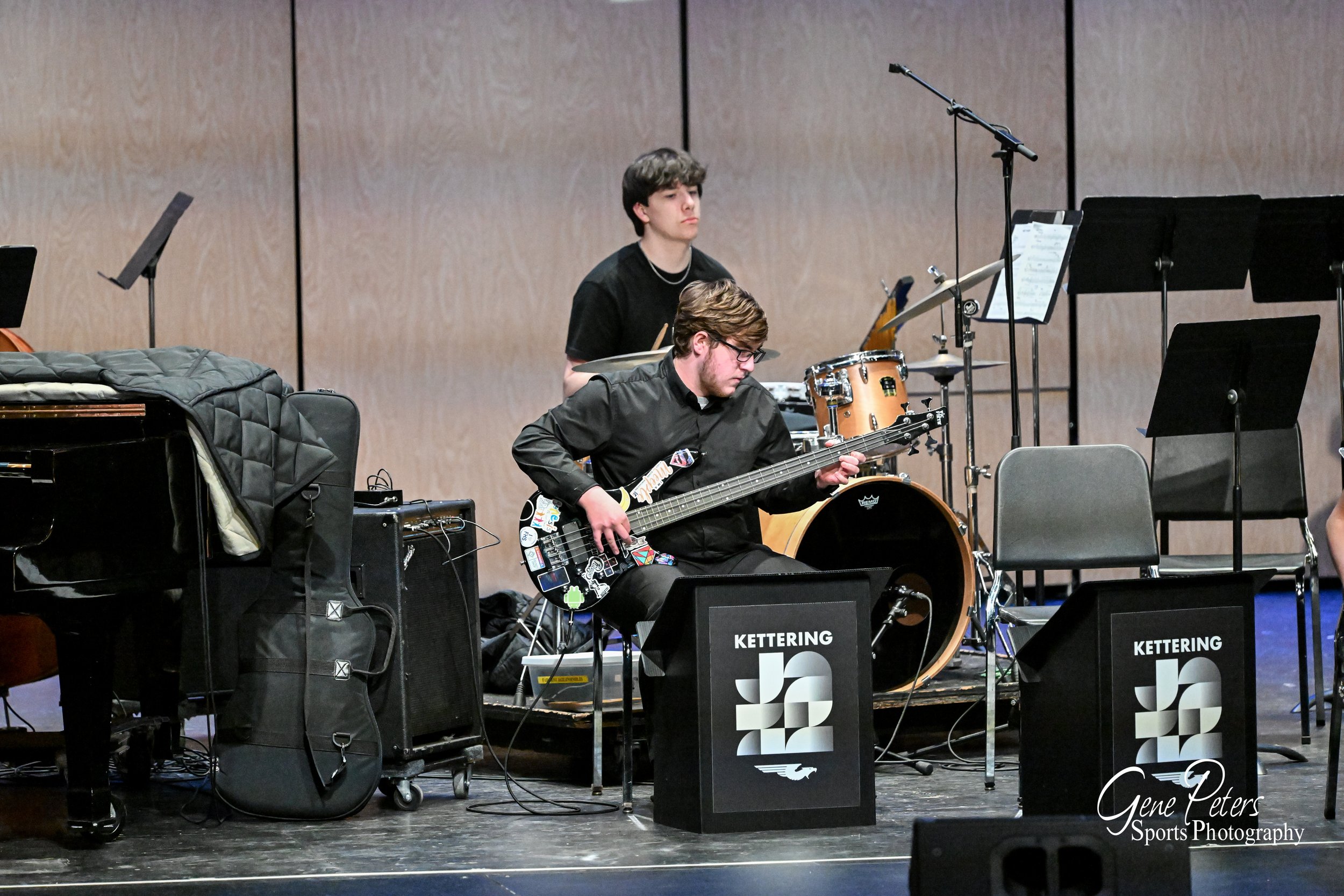 Musicians performing on stage with a piano, guitar, and drums, at Kettering Jazz event. Two young men are playing instruments, one on a bass guitar and the other on drums. Music stands and chairs are set up on stage.