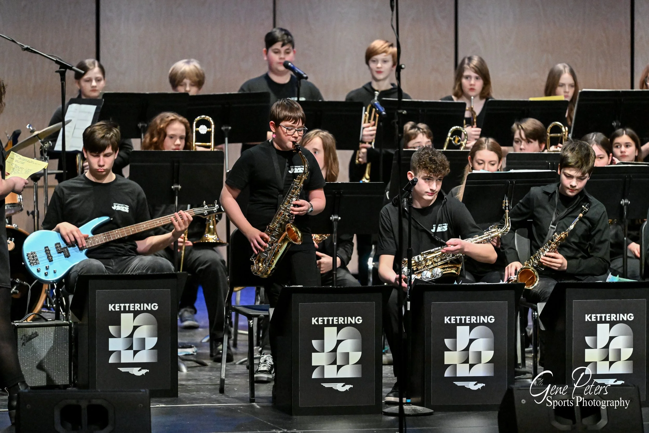 Young musicians in a school band performing on stage with guitars, saxophones, and music stands with 'Kettering Jazz' printed on front.