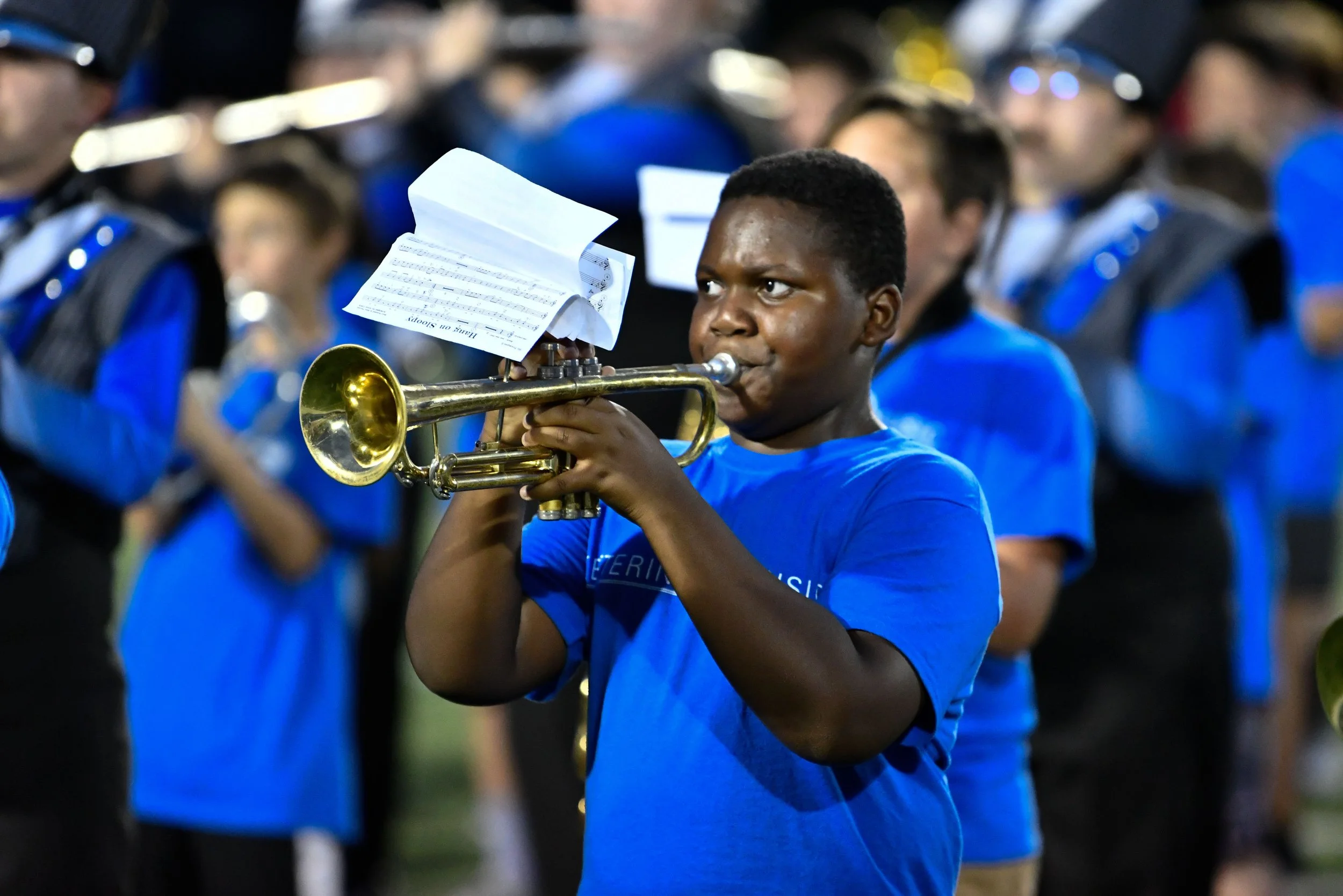 A young man wearing a blue shirt playing a trumpet during a marching band performance with other band members in blue and black uniforms in the background.