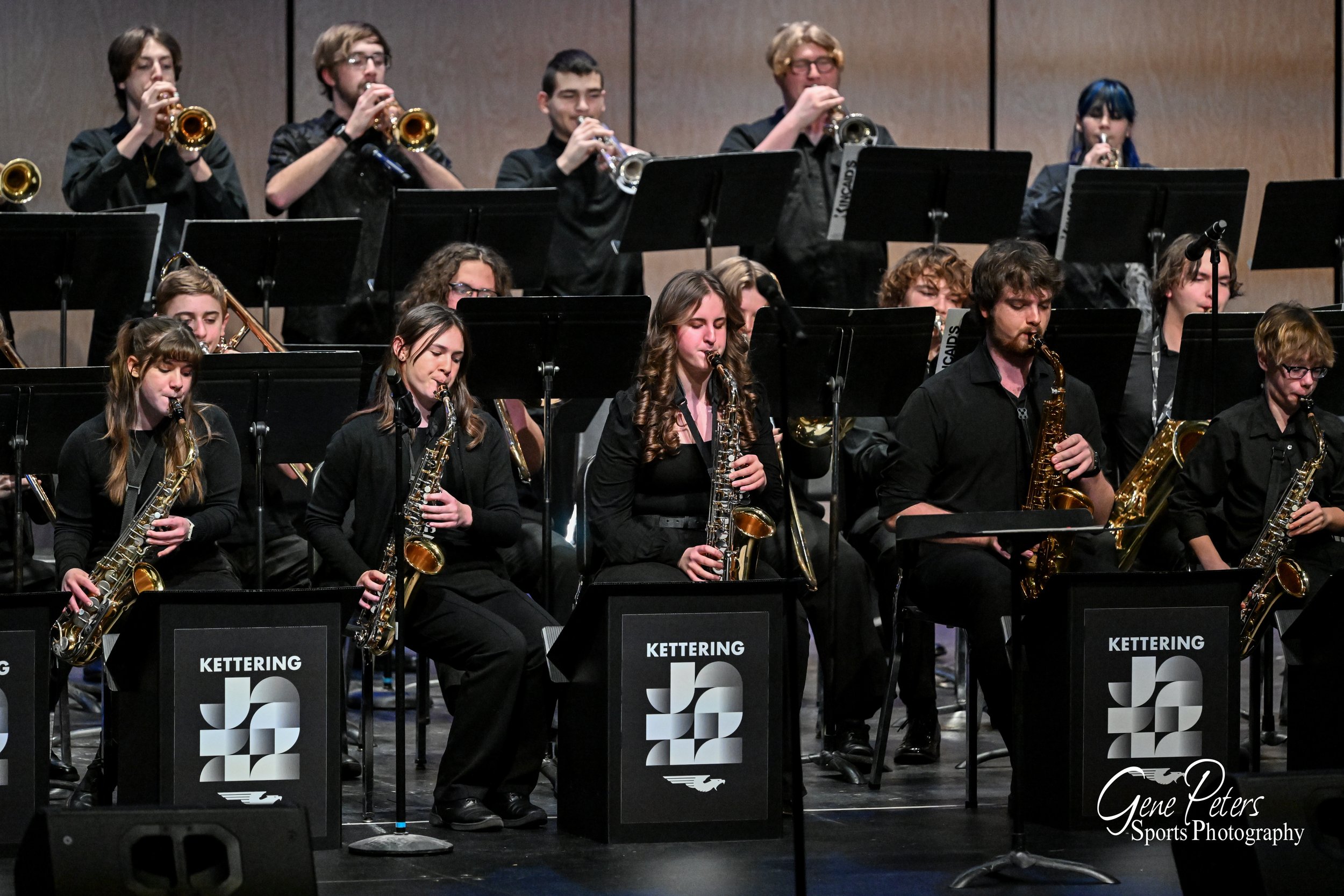 A large group of young musicians playing saxophones and trumpets on stage at a concert, with music stands displaying 'Kettering' and a logo, under stage lighting.