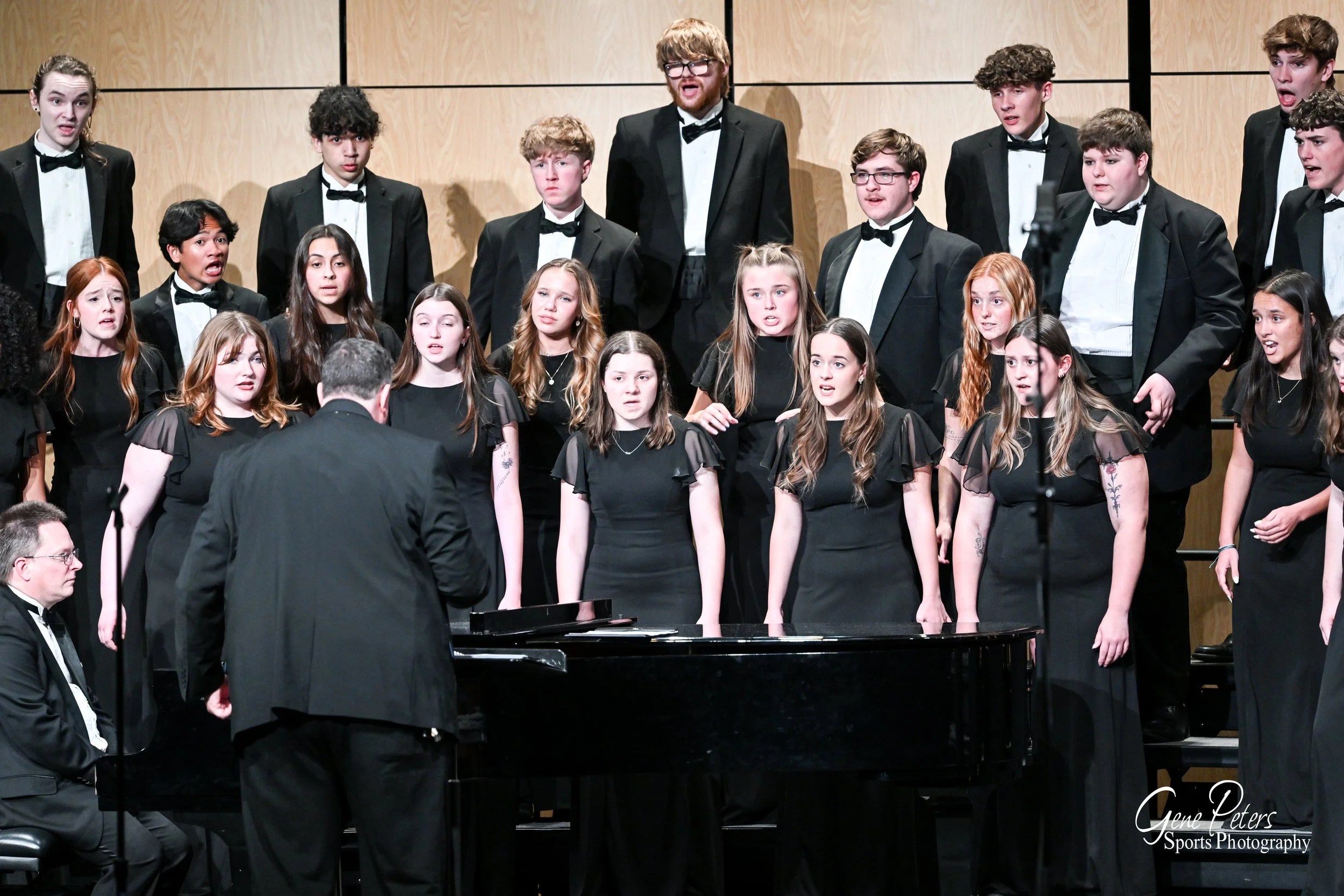 A choir performing on stage, directed by a conductor, with a pianist in front of them, all dressed in black formal attire.