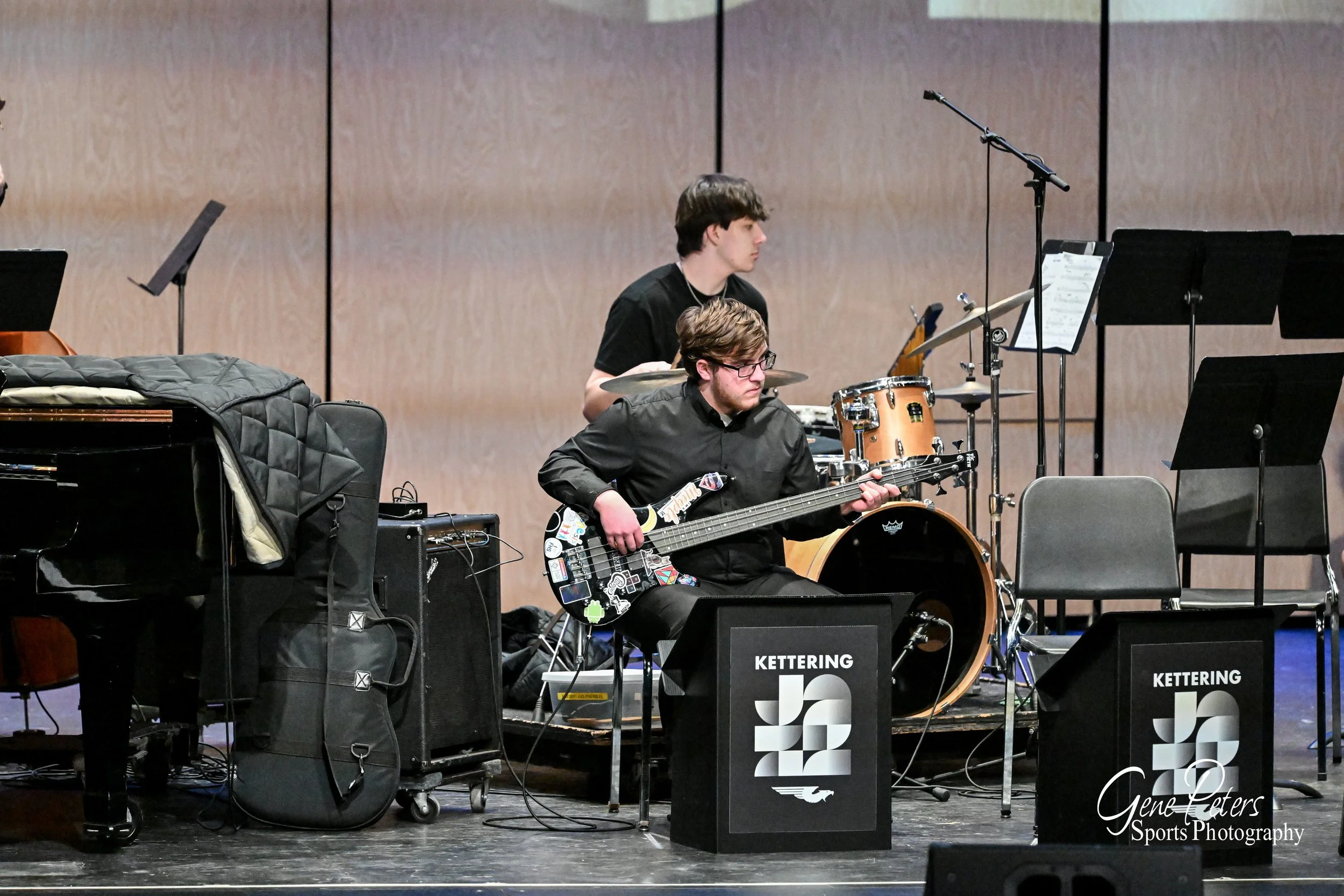 Two young men playing musical instruments on a stage; one playing an electric guitar, the other on drums. There are music stands and black speakers with the Kettering Jazz logo.