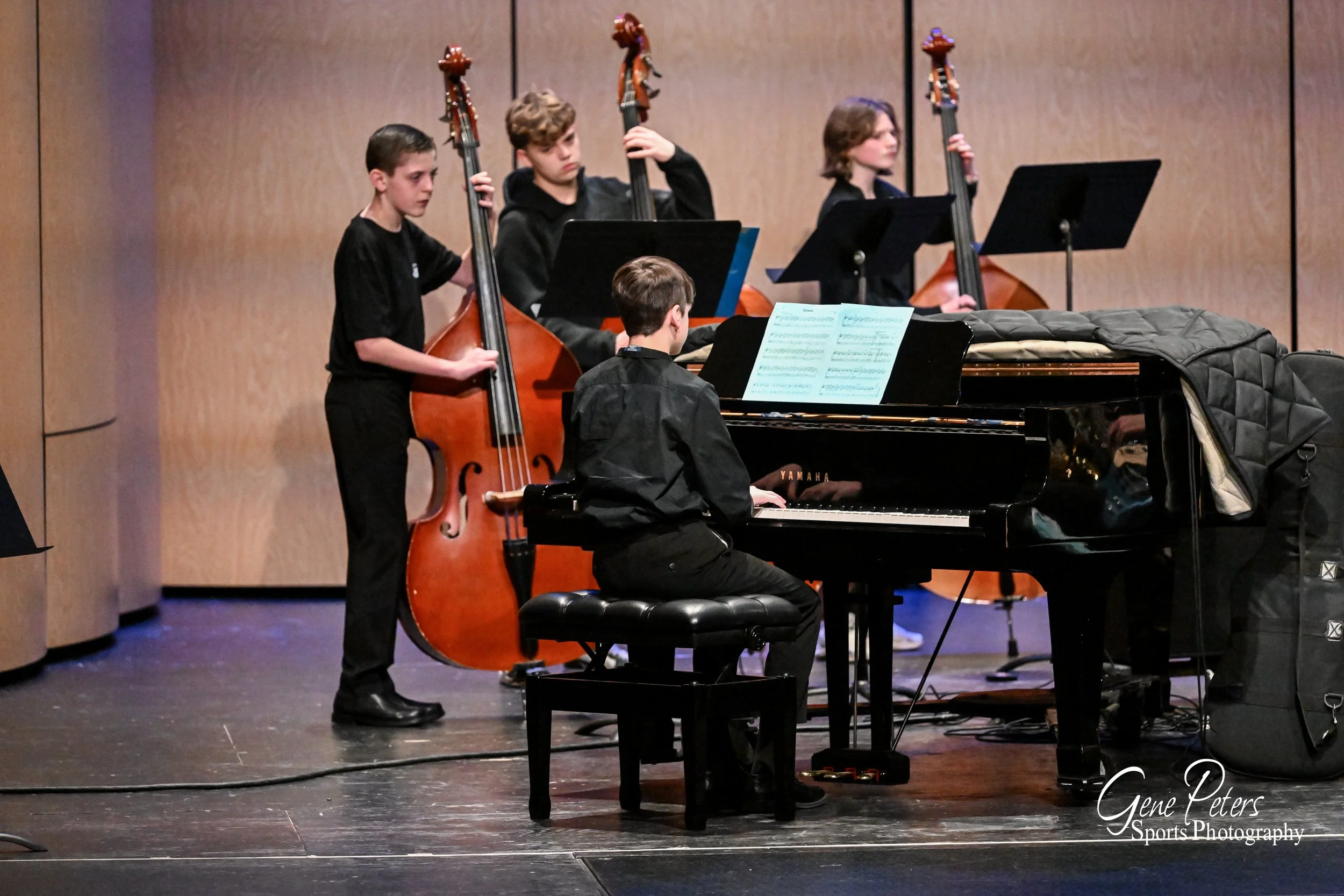 A group of young boys playing musical instruments during a performance. One boy is playing a grand piano, while three others are playing double basses in the background. The scene is set on stage with a wooden wall backdrop.