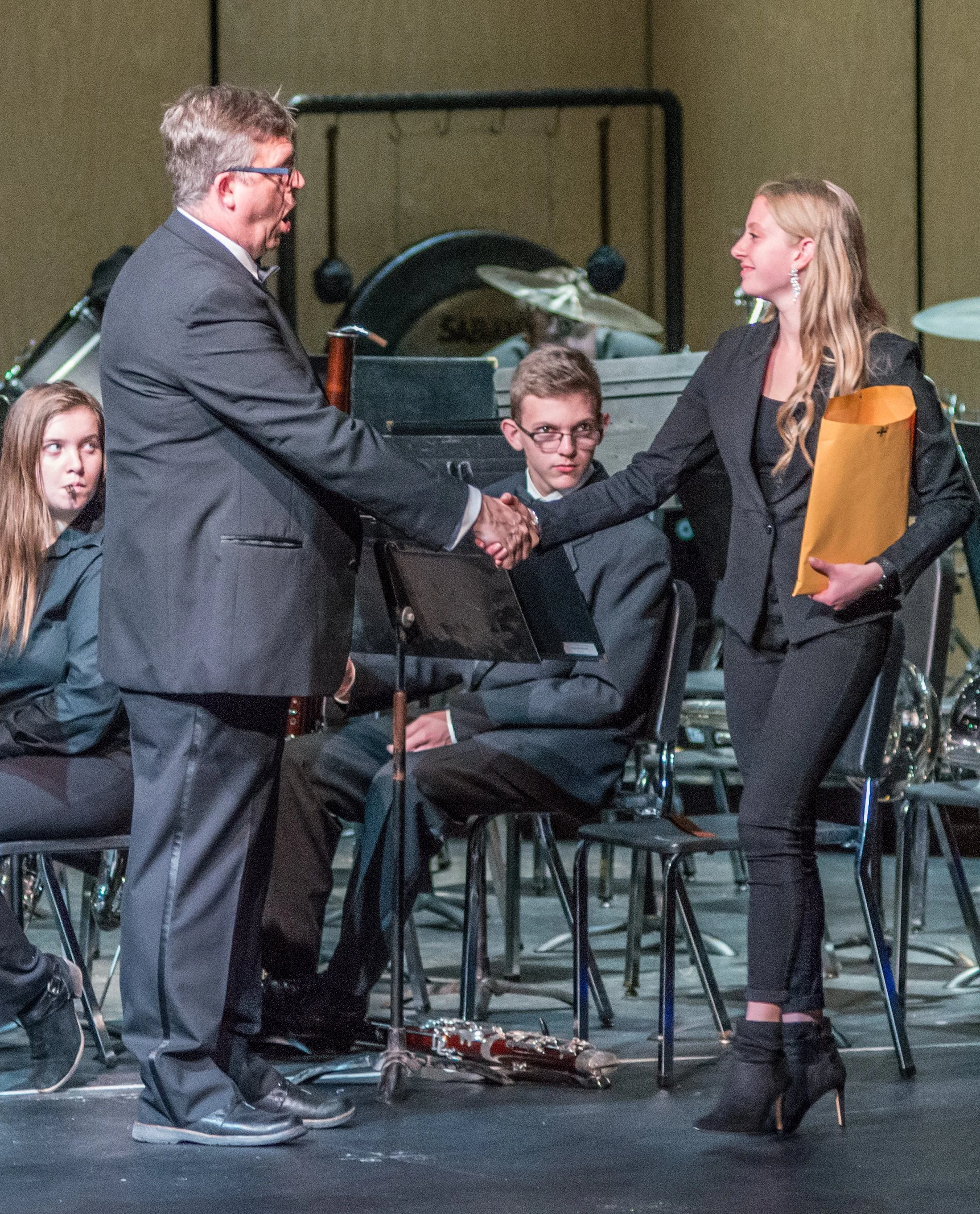 A woman in black heels and a man in glasses shake hands during a formal event, with seated musicians in the background.