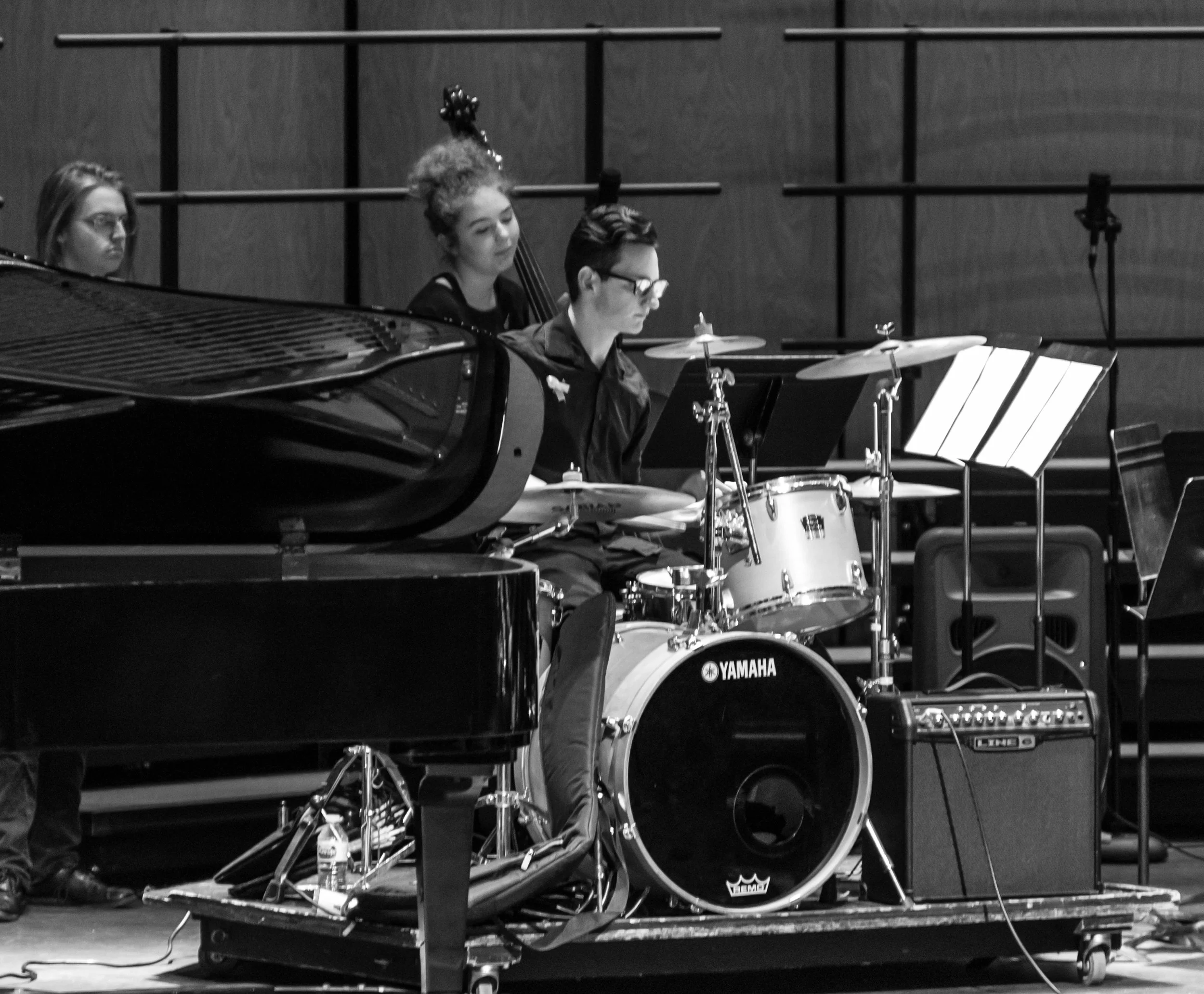 Black and white photo of musicians performing on stage, featuring a grand piano, drums, a double bass, and music stands, with a wooden wall backdrop.