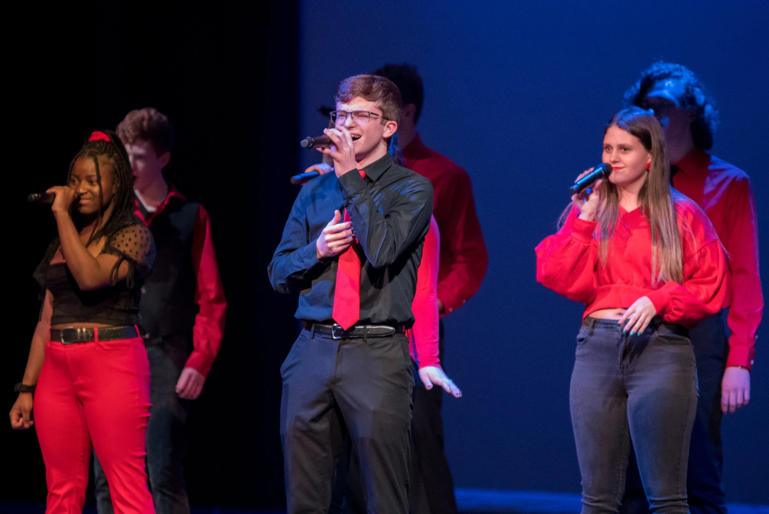 A group of young people performing on stage, with two holding microphones. They are dressed in red, black, and dark clothing, standing against a dark backdrop.