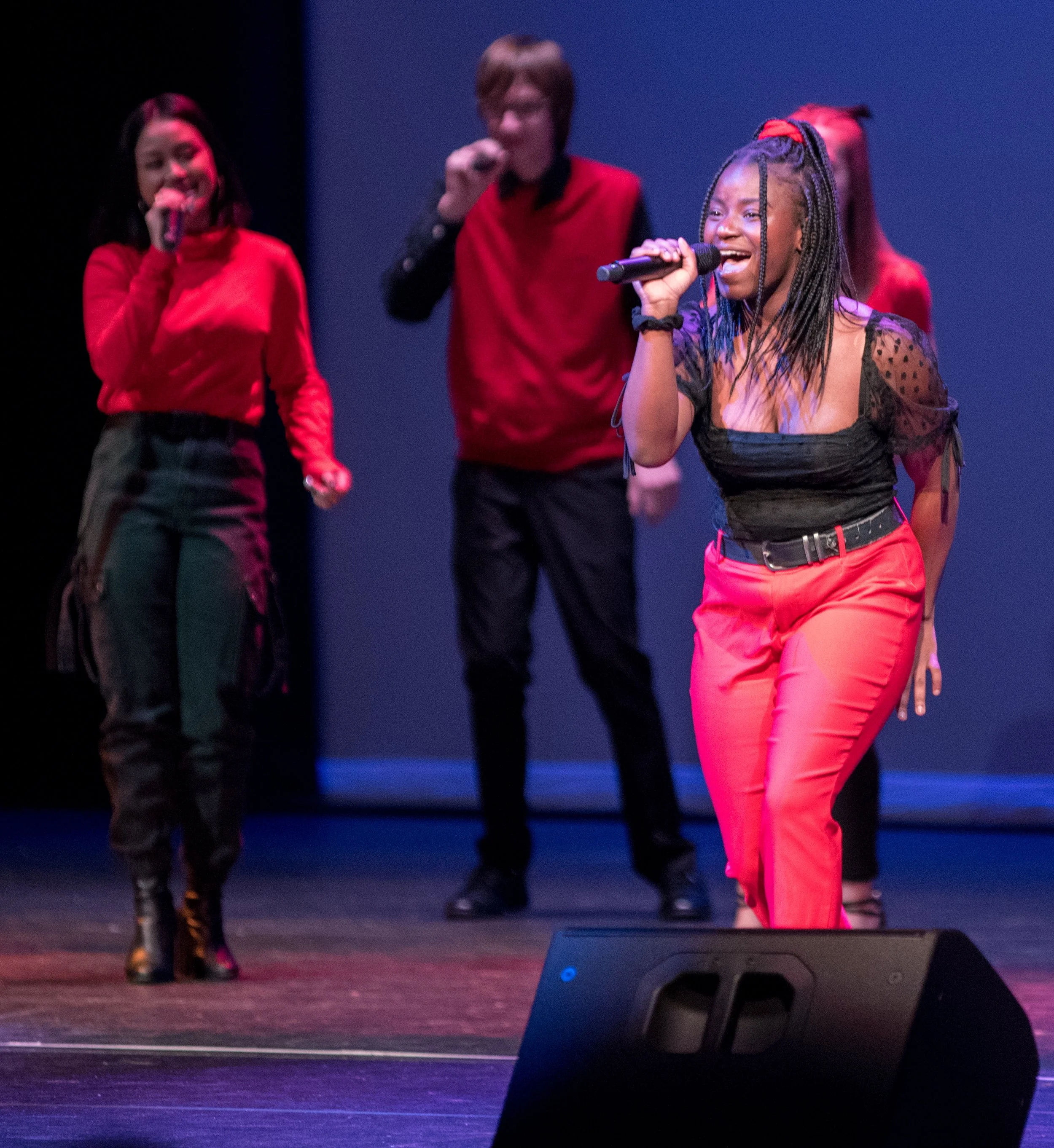 Four people performing on stage, three women holding microphones and singing, one man in the background, all dressed in red and black outfits.