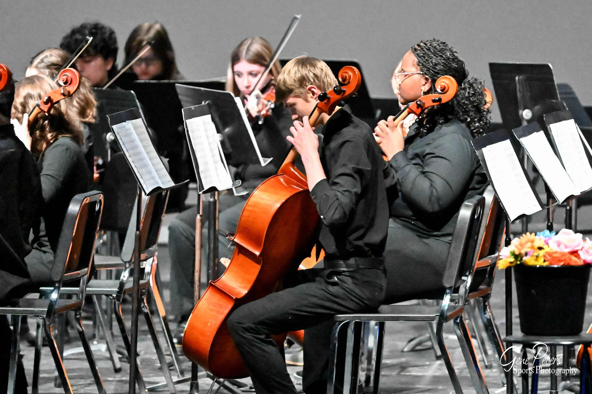 Group of young musicians in black formal attire playing violins and a cello during a concert on stage, with music stands and sheet music in front of them.