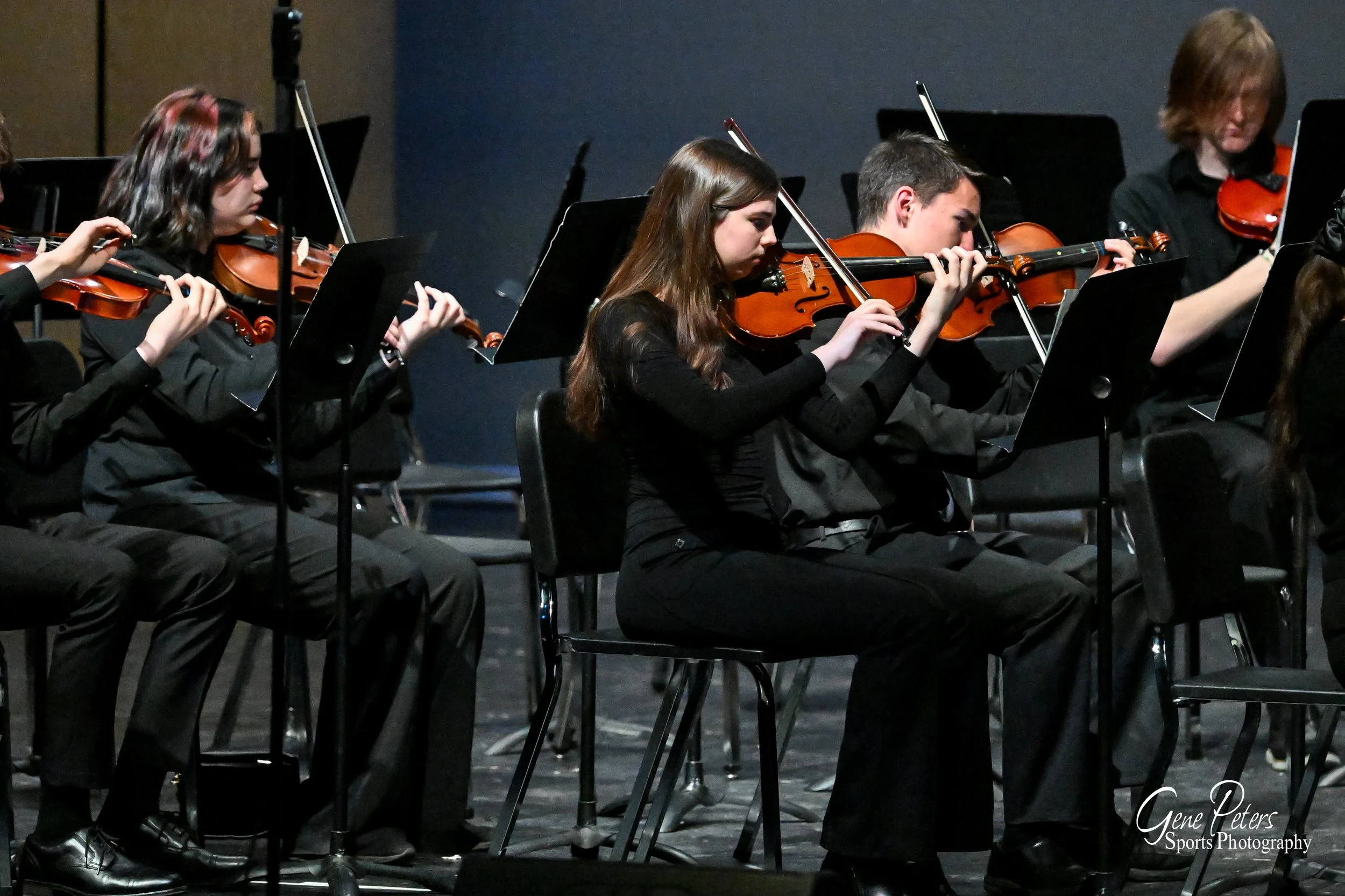 Young musicians playing violins during a performance on stage, dressed in black, with music stands in front of them.