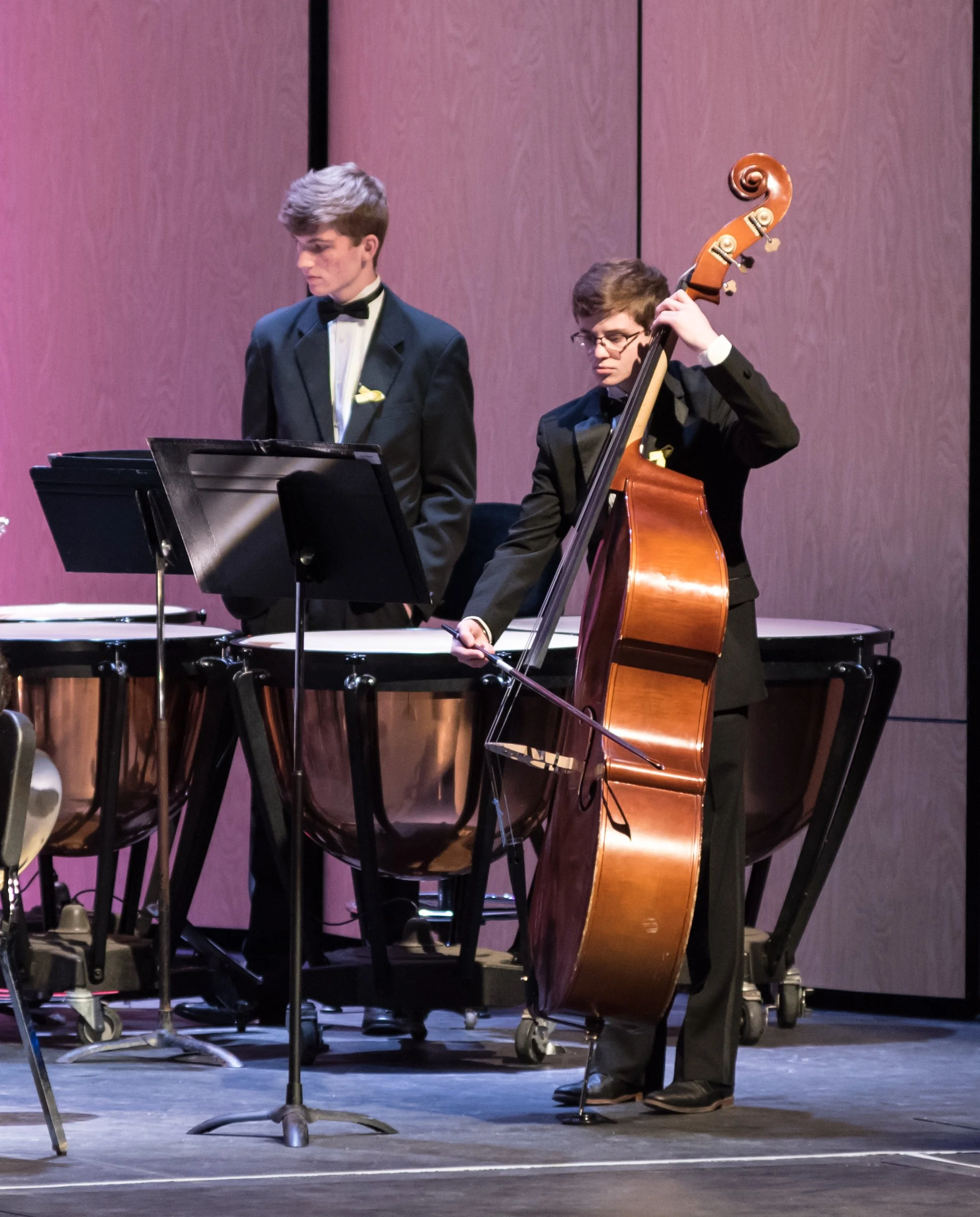 Two young men in tuxedos performing music on stage, one playing a double bass and the other on a percussion instrument, with music stands in front of them.