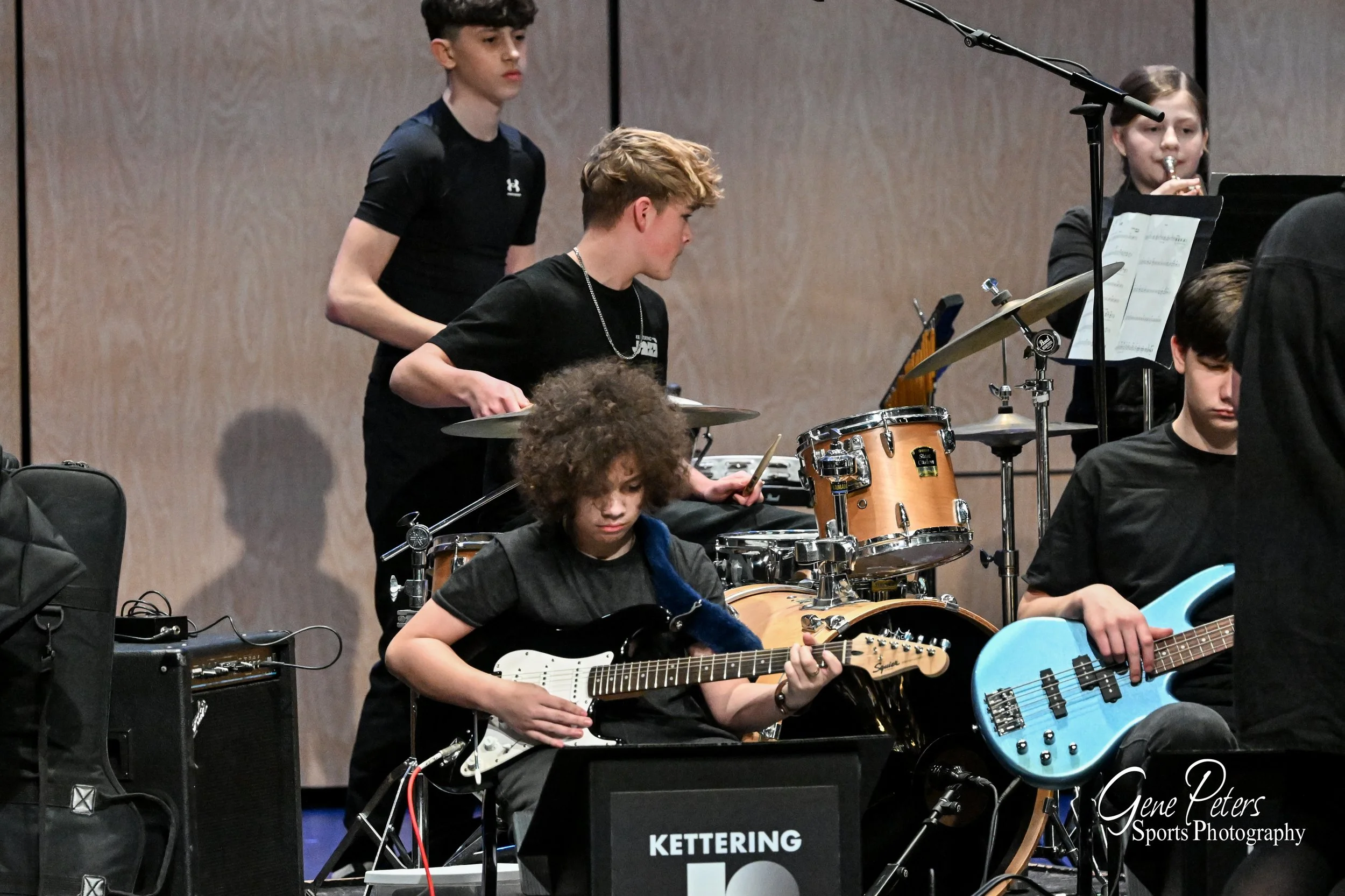 Young students playing musical instruments in a band, including electric guitar, bass guitar, drums, and woodwind, during a performance or rehearsal.