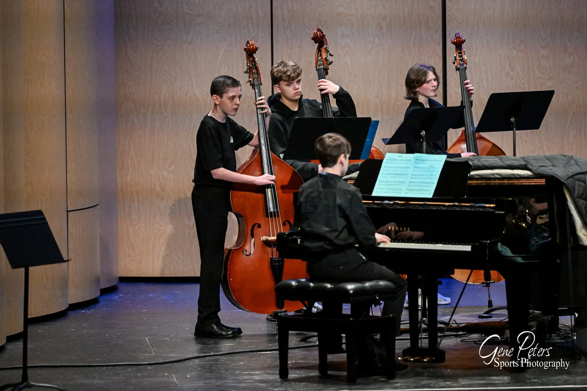 Four young musicians playing a piano and upright bass on stage, with sheet music on stands, in a concert hall with wooden panels.