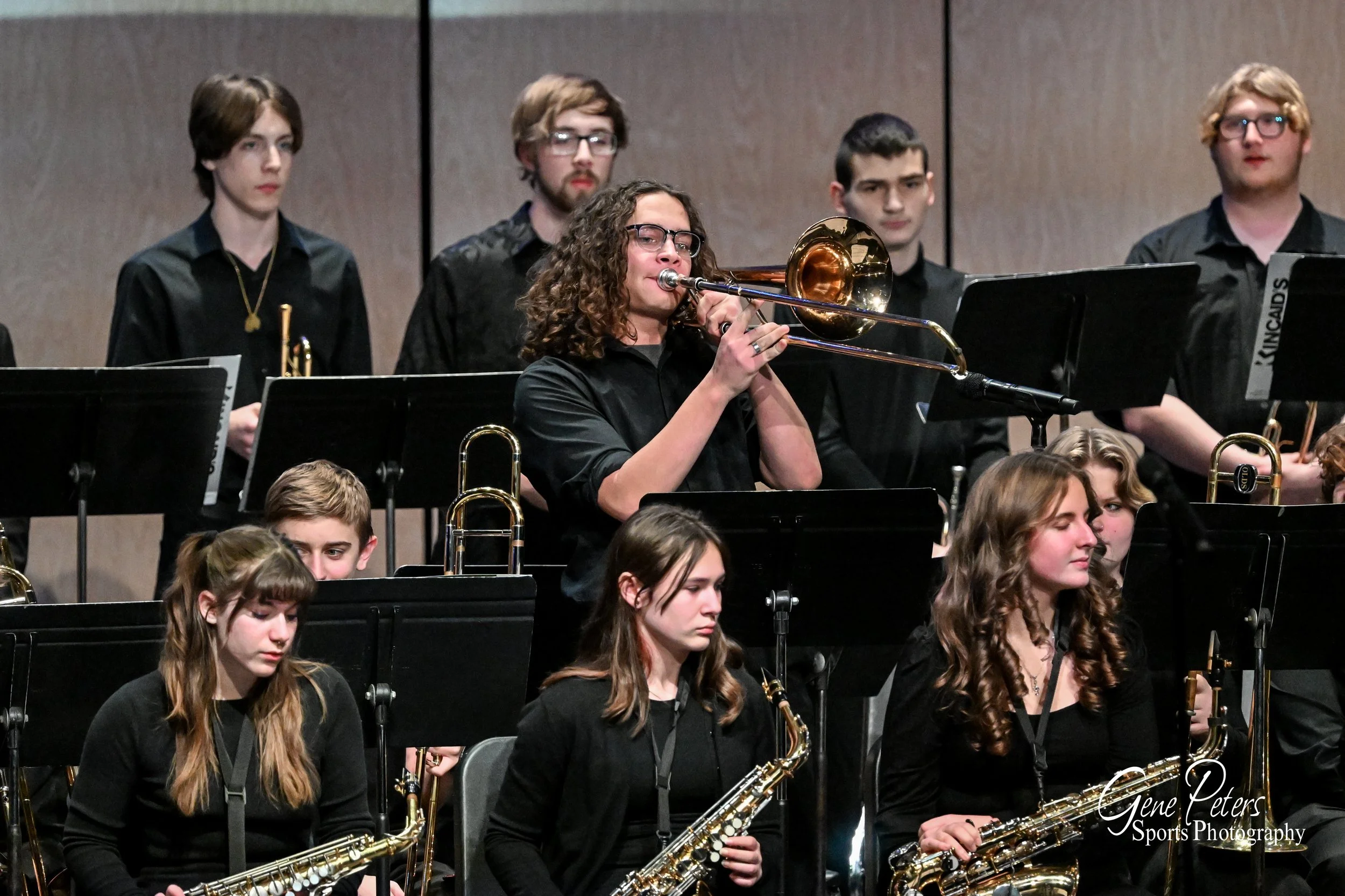 A group of young musicians in black clothing performing in an orchestra, with a female musician playing the trombone in the center.