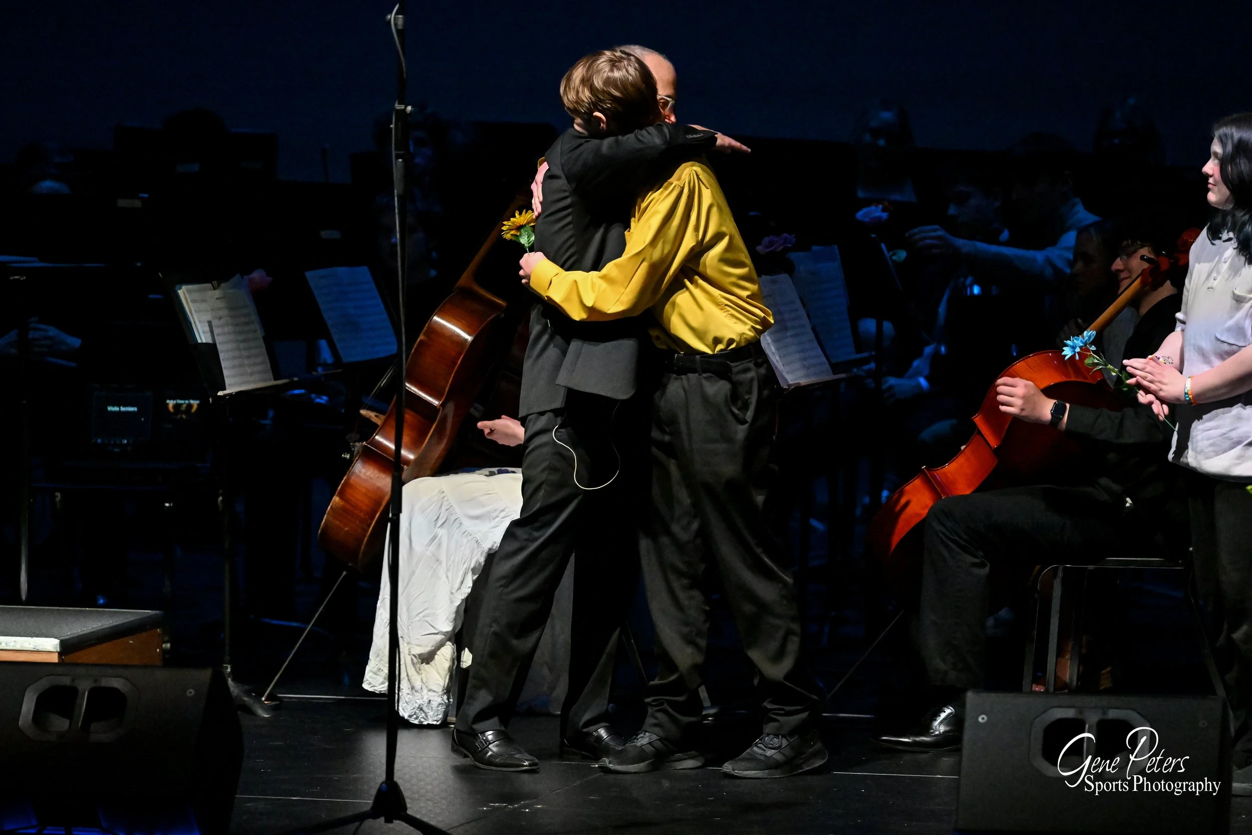 Two people hugging on stage, with a seated musician holding a cello nearby, in a dark concert hall setting.