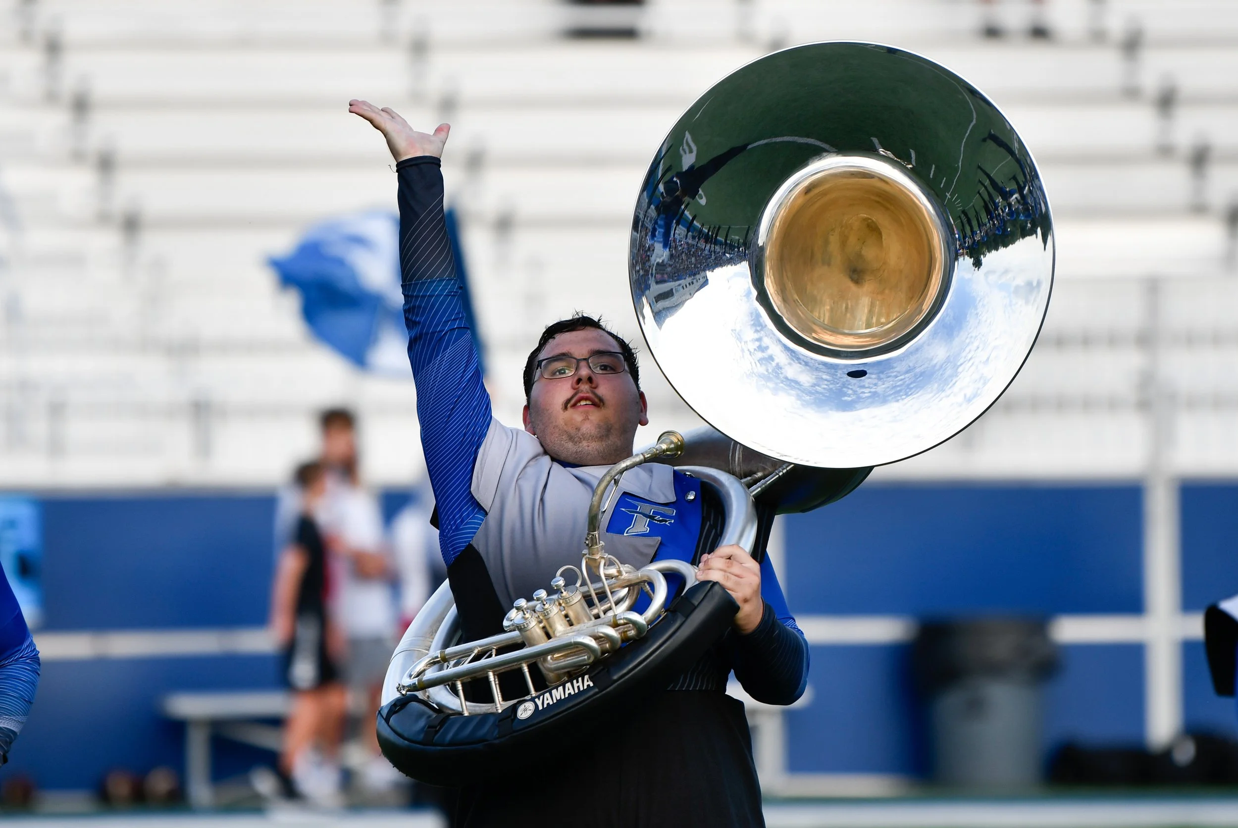 A man holding a sousaphone at a sports stadium, with a raised arm and a flag in the background.