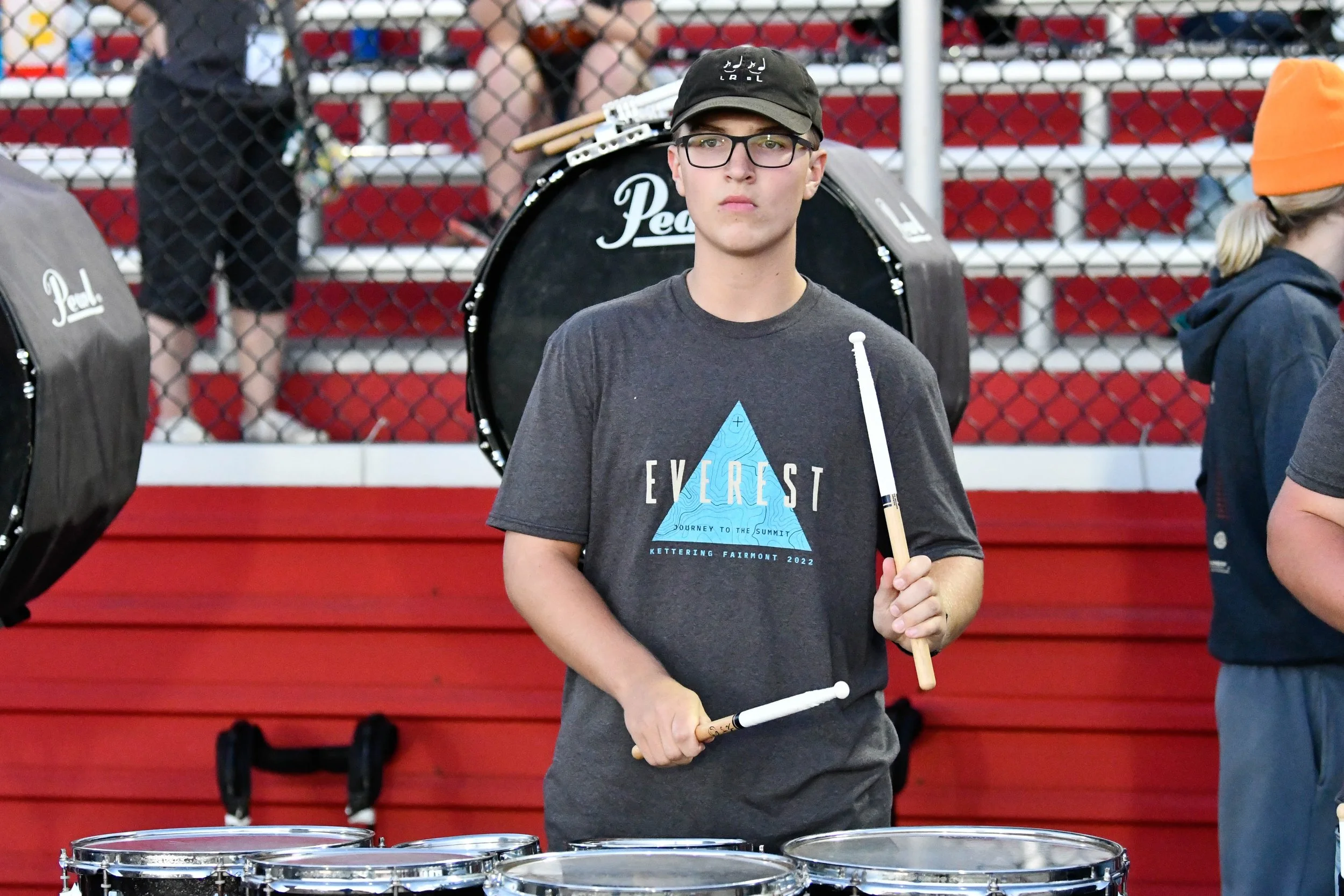 A teenage boy in a dark grey t-shirt with the word 'Everest' and a mountain graphic printed on it, wearing glasses and a black cap, holding a drumstick in each hand, standing in front of a row of drums on a red-painted bleacher, with other people aro