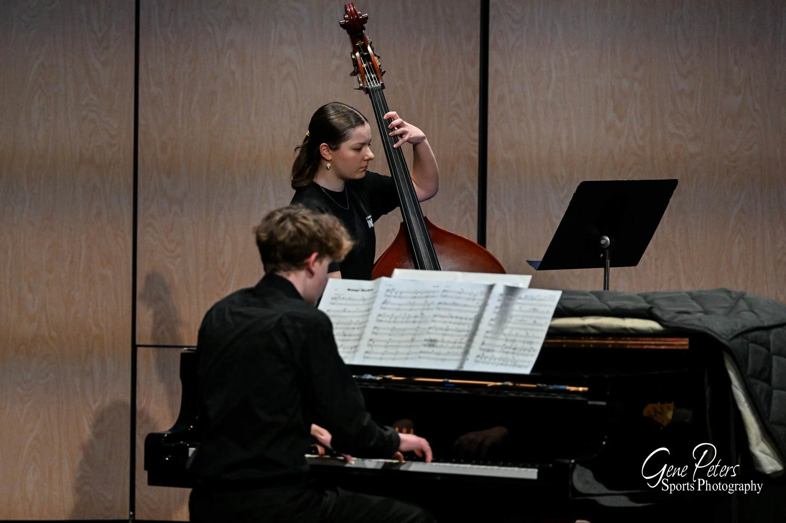 Two musicians, one playing the piano with sheet music and the other playing the double bass, during a performance in a room with wood-paneled walls.