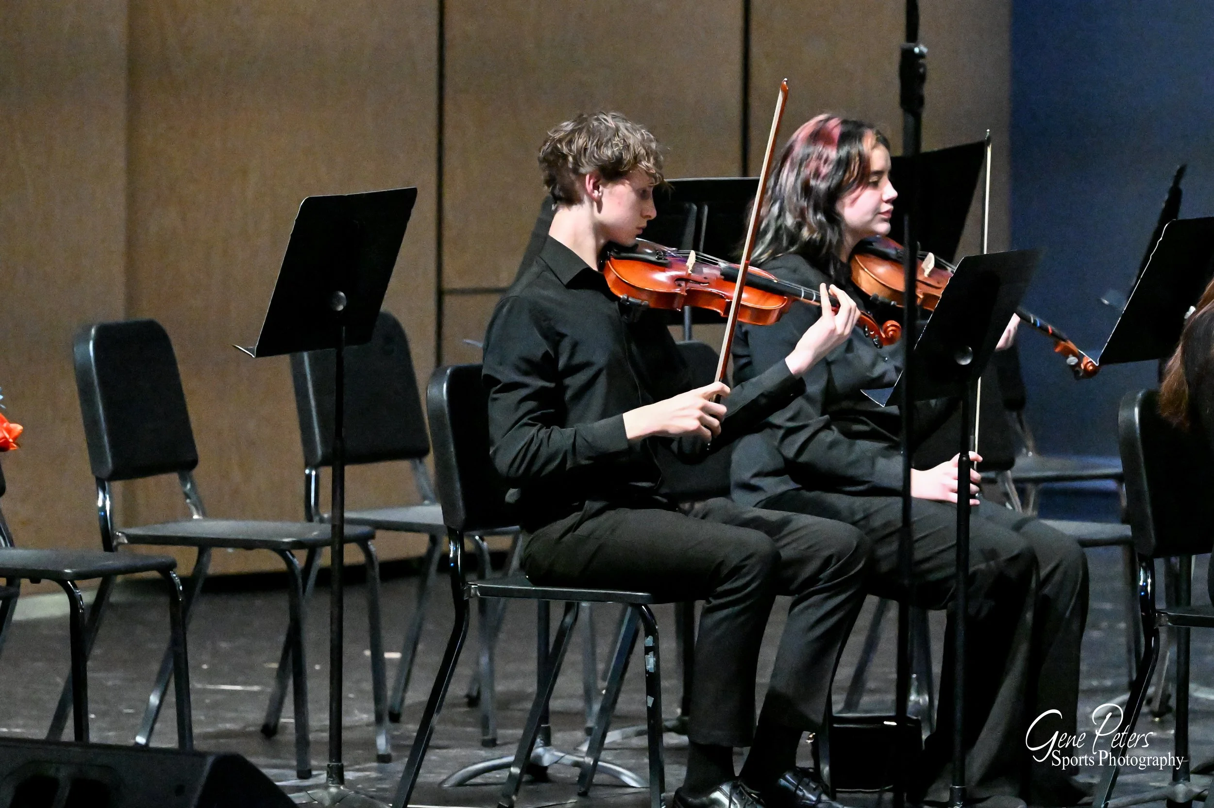 Two young musicians, a boy and a girl, dressed in black, sitting on chairs and playing violins during a concert. They are focused on their music, with music stands in front of them and a stage background.