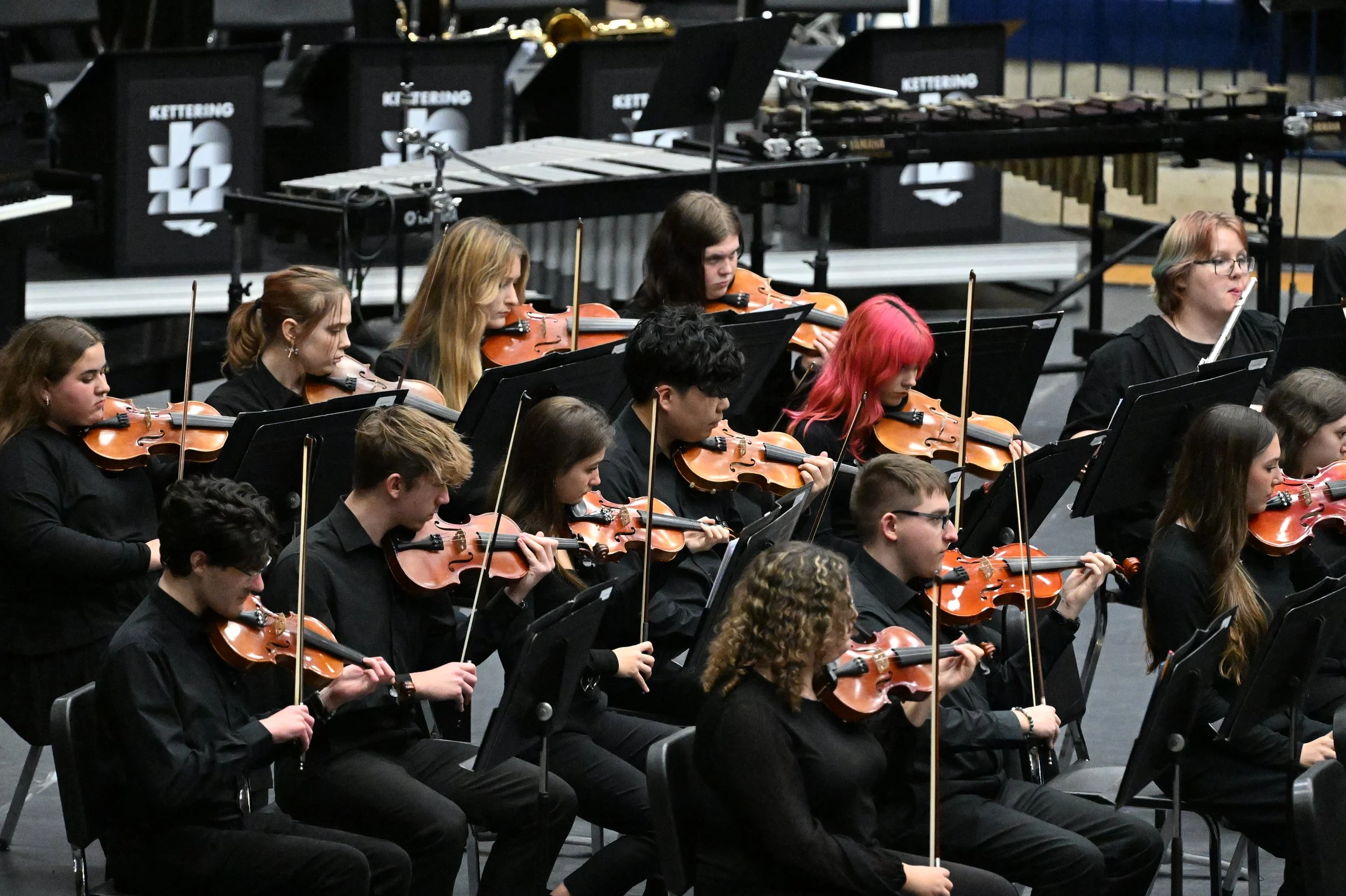 Orchestra of young musicians playing violins during a performance, with music stands and part of a percussion setup in the background.