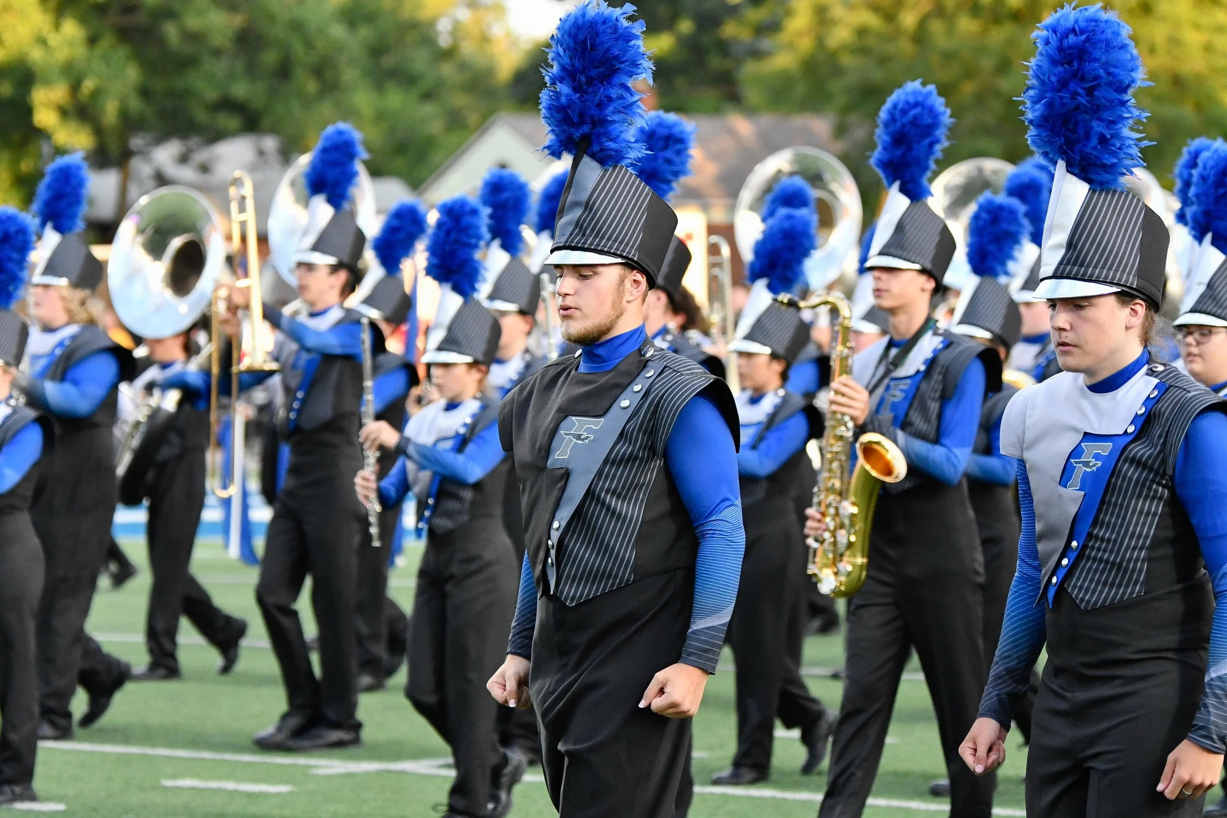 A marching band performing on a football field, with members dressed in black and blue uniforms and tall black hats with blue feather plumes, playing various brass and woodwind instruments.
