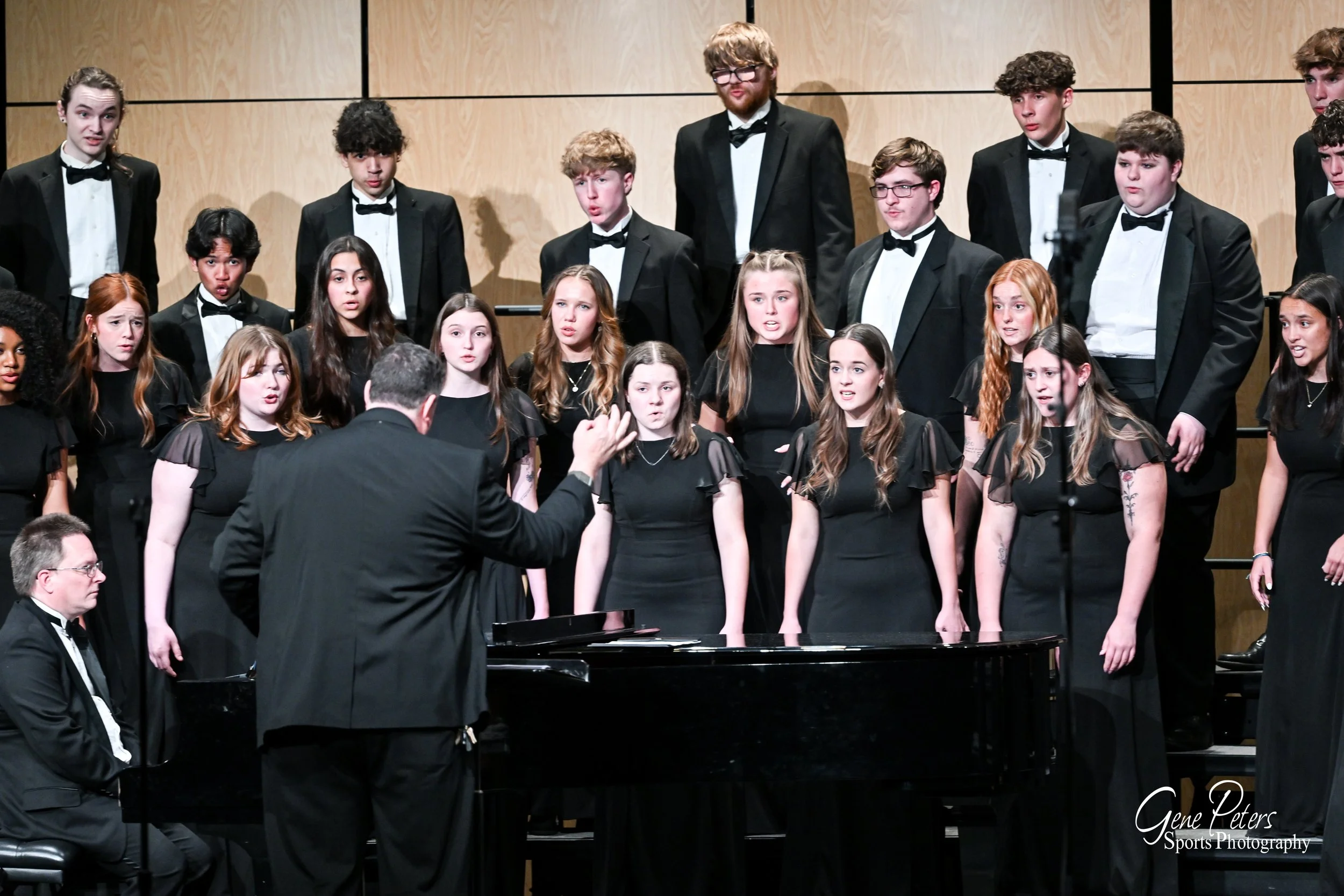 A choir performing on stage, directed by a conductor, with a pianist seated at a grand piano.