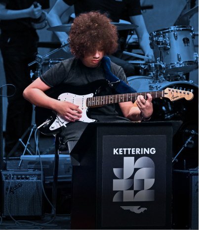 Young person with curly hair playing an electric guitar at Kettering event.