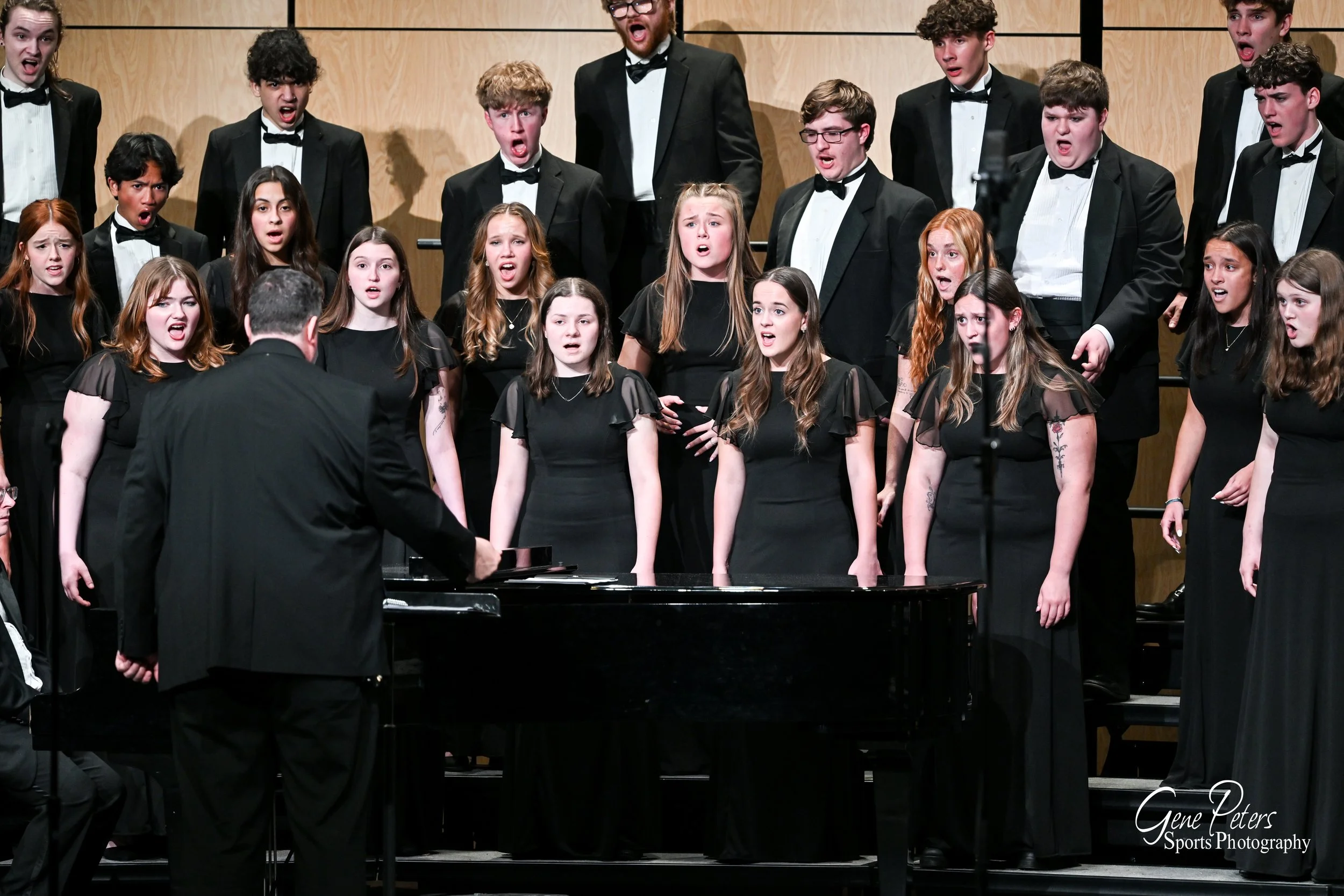 A choir of young men and women singing on stage, conducted by a man in a tuxedo, with a grand piano in front. The choir members are dressed in formal black attire.