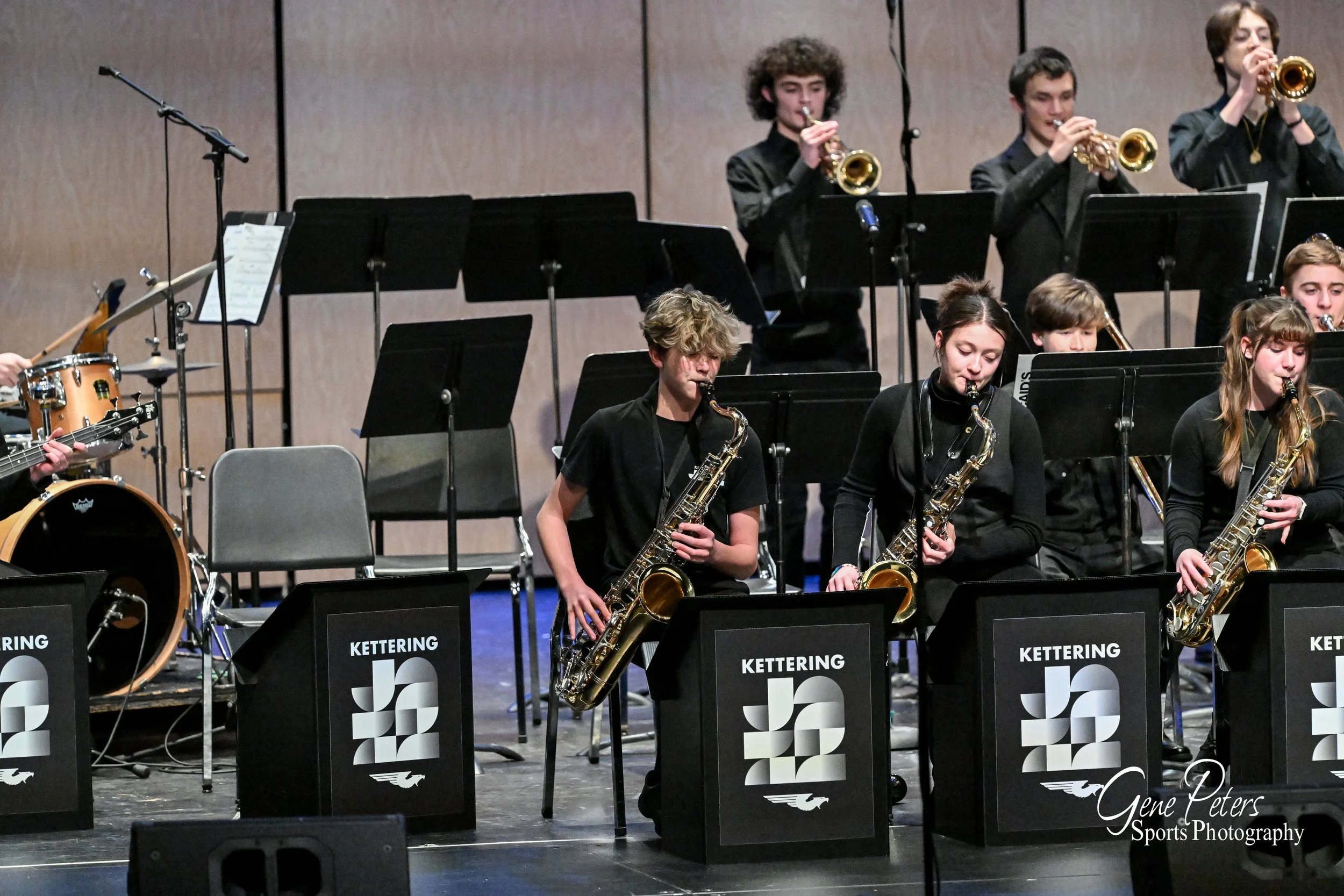 Young musicians playing saxophones and trumpet during a concert on stage, with music stands labeled 'Kettering Jazz' and a drum set in the background.