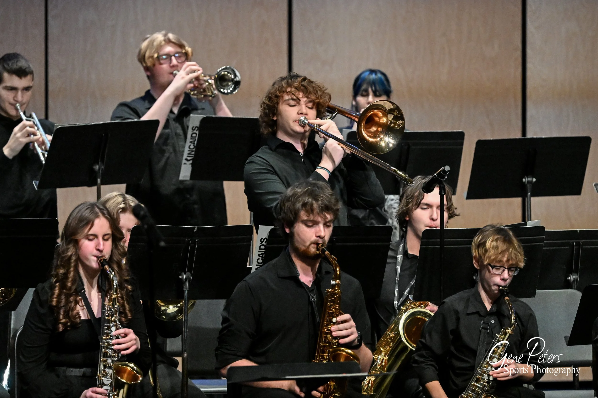 Group of young musicians performing in an orchestra, playing various wind instruments, dressed in black, with a wood-paneled wall background.