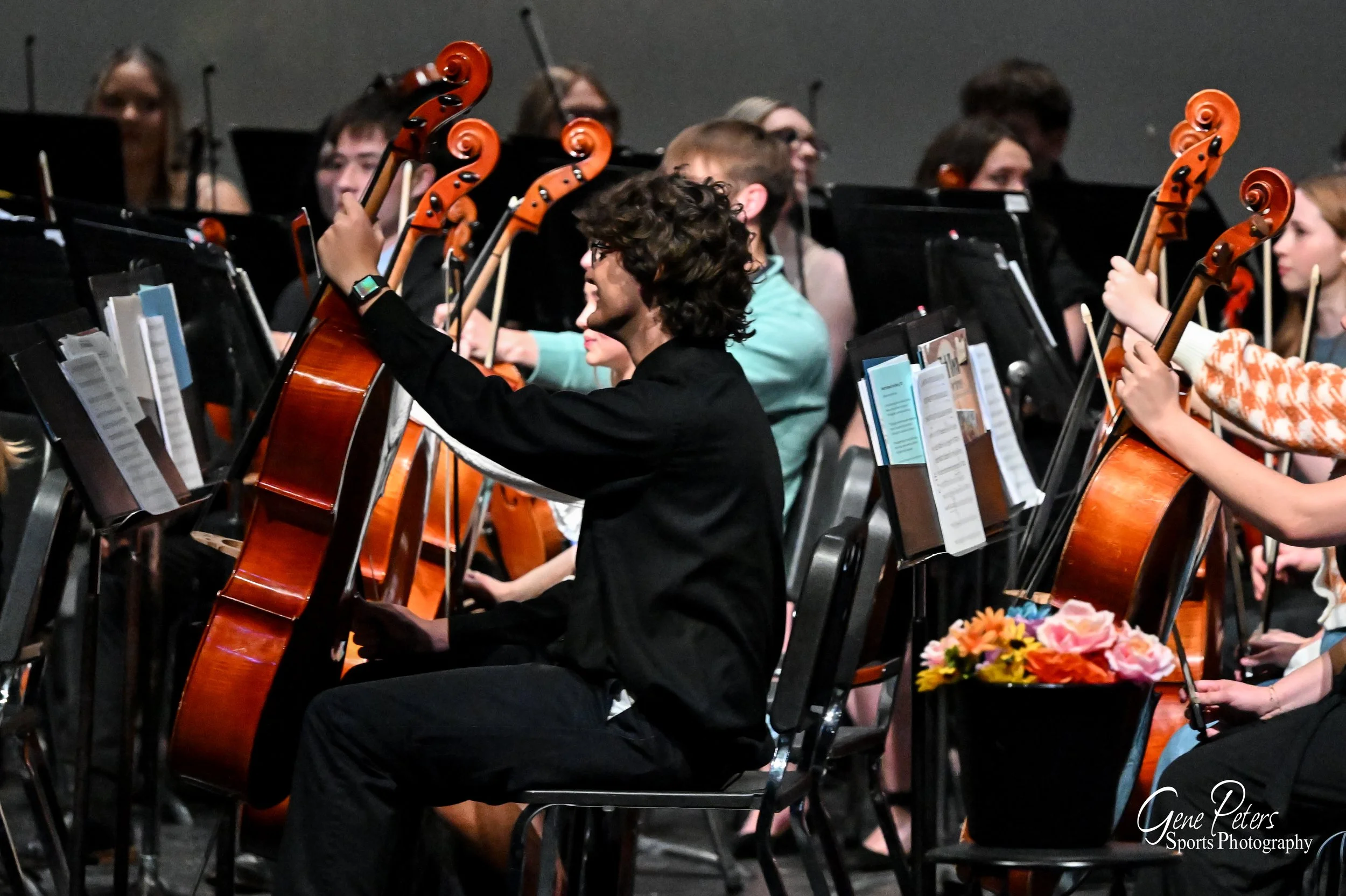 Young orchestra musicians playing cellos during a performance, with sheet music and a flower arrangement on a chair in the foreground.