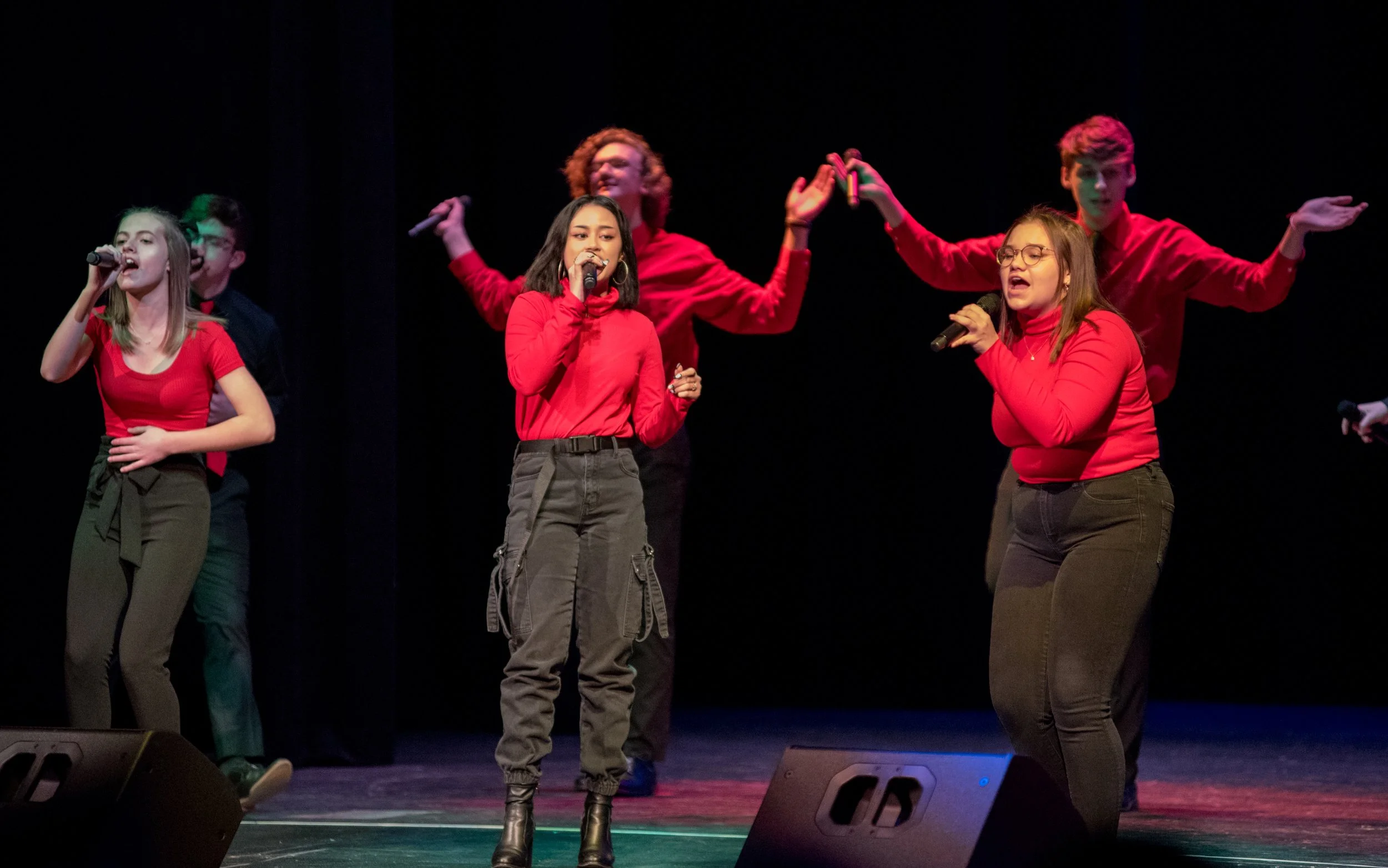 Six young performers singing on stage, all wearing red tops and dark pants, with black curtains in the background.