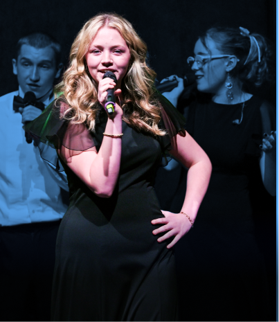 Young woman singing into a microphone on stage with two people in the background.