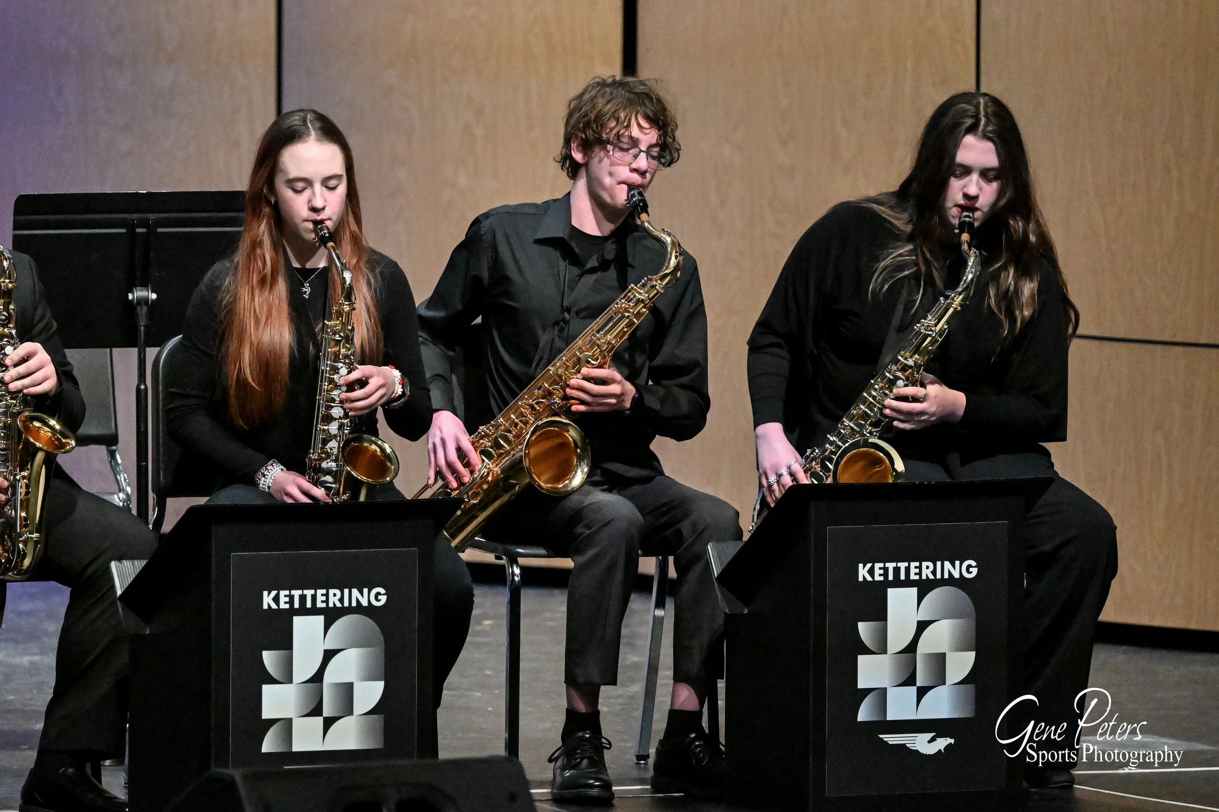 High school jazz band performing on stage, featuring three students with saxophones, wearing black attire, with music stands labeled 'Kettering' in front.