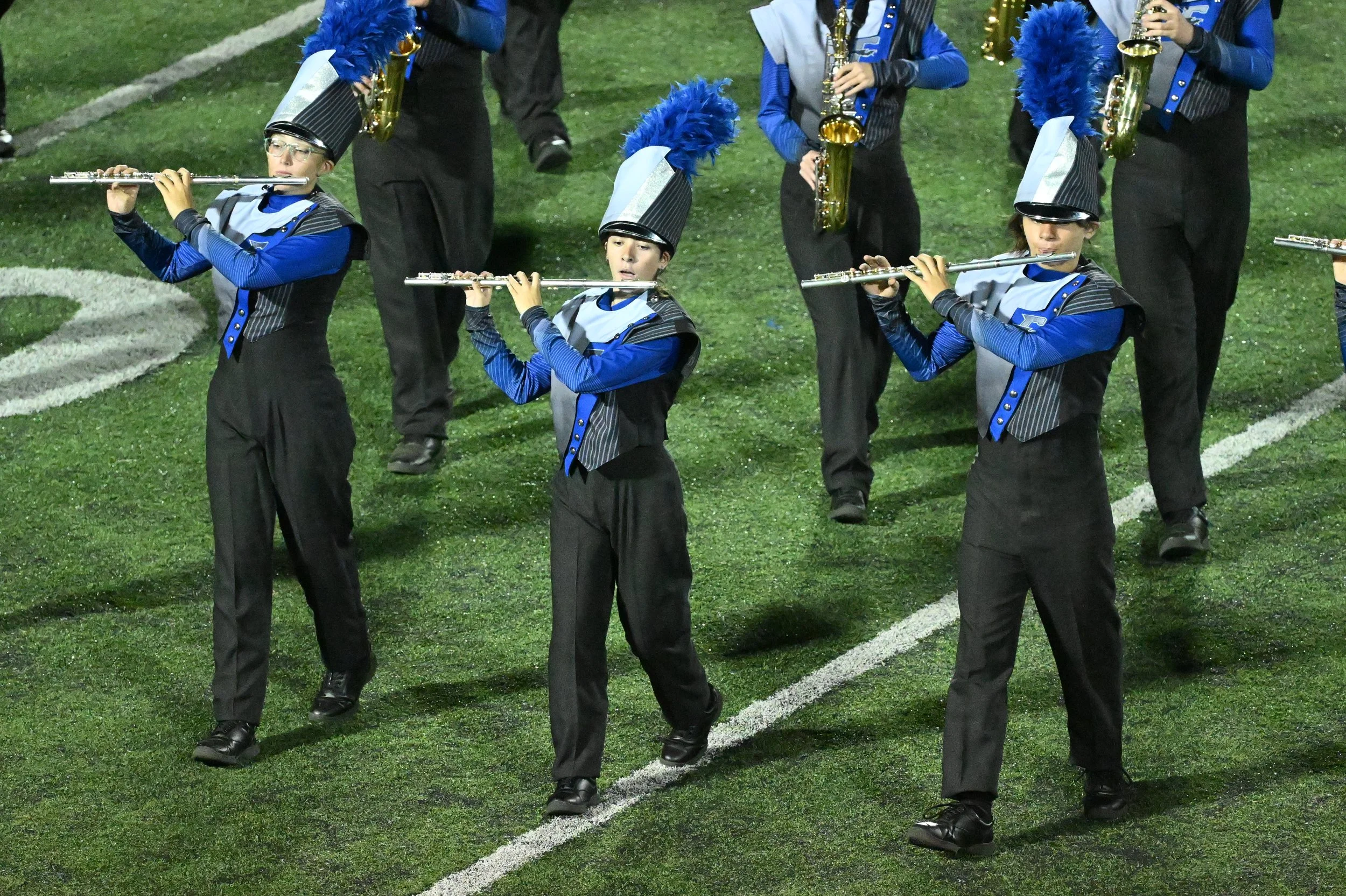 A group of young marching band members playing flutes during a performance on a football field. They are dressed in blue and black uniforms with tall black hats adorned with blue feathers.