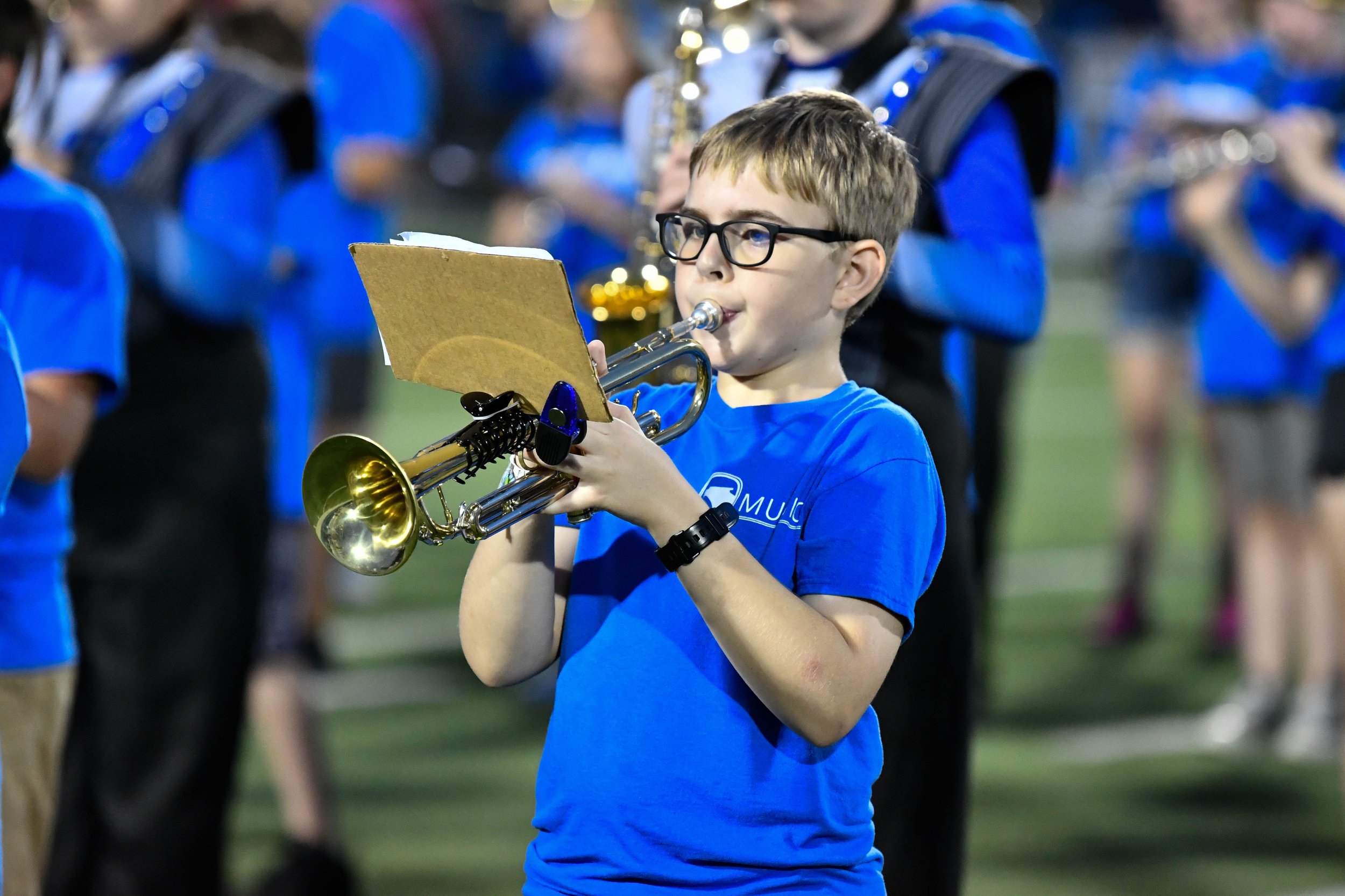Young boy wearing glasses and a blue shirt playing the trumpet at a band performance on a field, with other band members in similar blue shirts in the background.